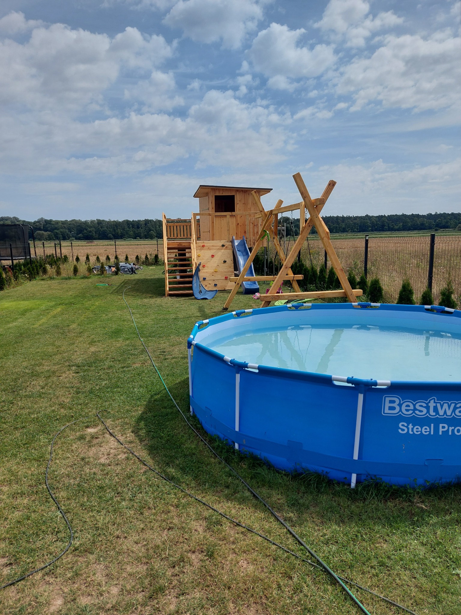 Plac zabaw z drewnianym domkiem, zjeżdżalnią, ścianką wspinaczkową i huśtawką obok rozłożonego basenu Bestway Steel Pro na trawniku w ogrodzie.