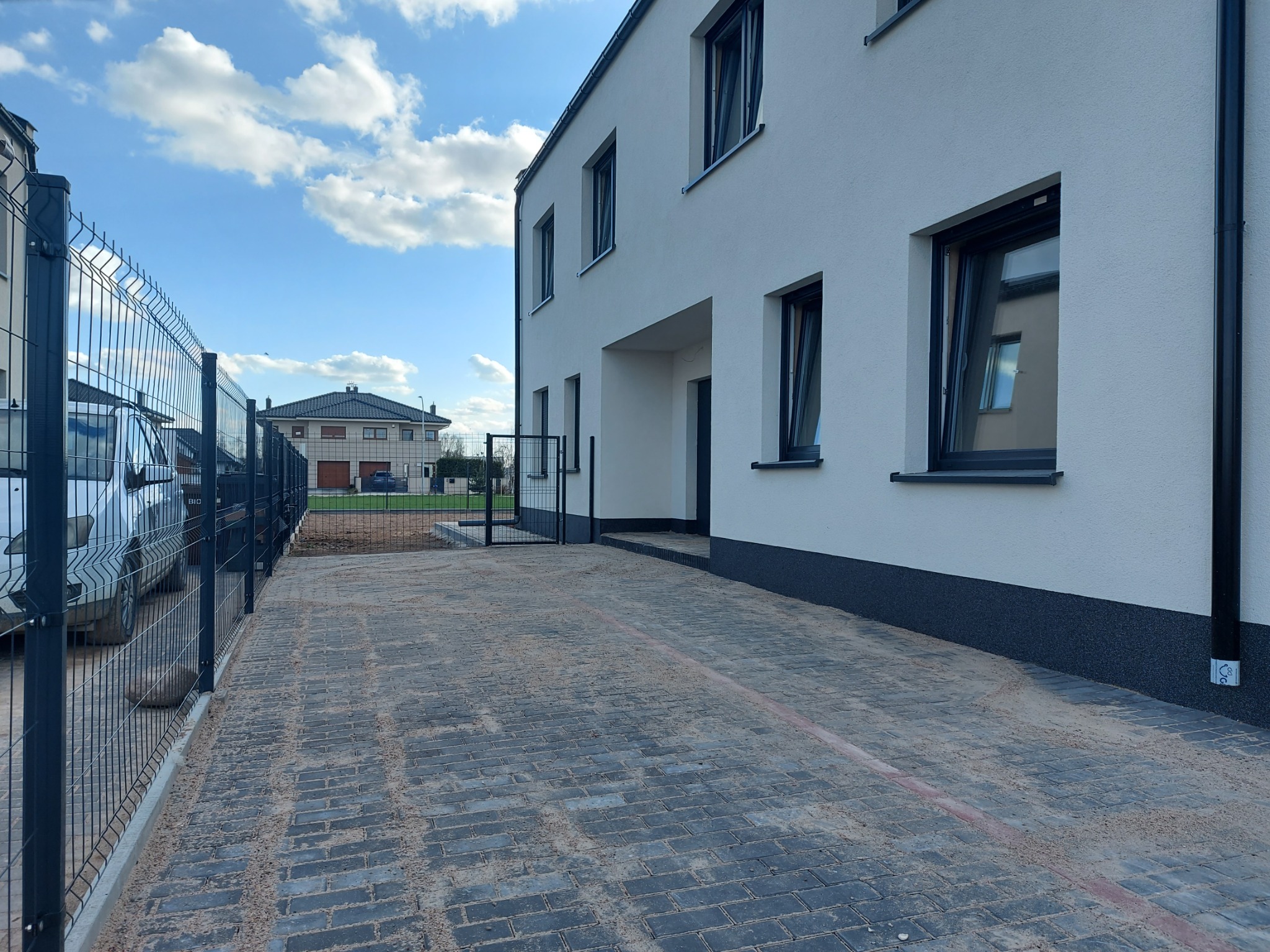 Nowoczesny, dwukondygnacyjny dom z białym tynkiem, czarnymi ramami okiennymi i brukowanym podjazdem, widoczny przez metalowe ogrodzenie panelowe.