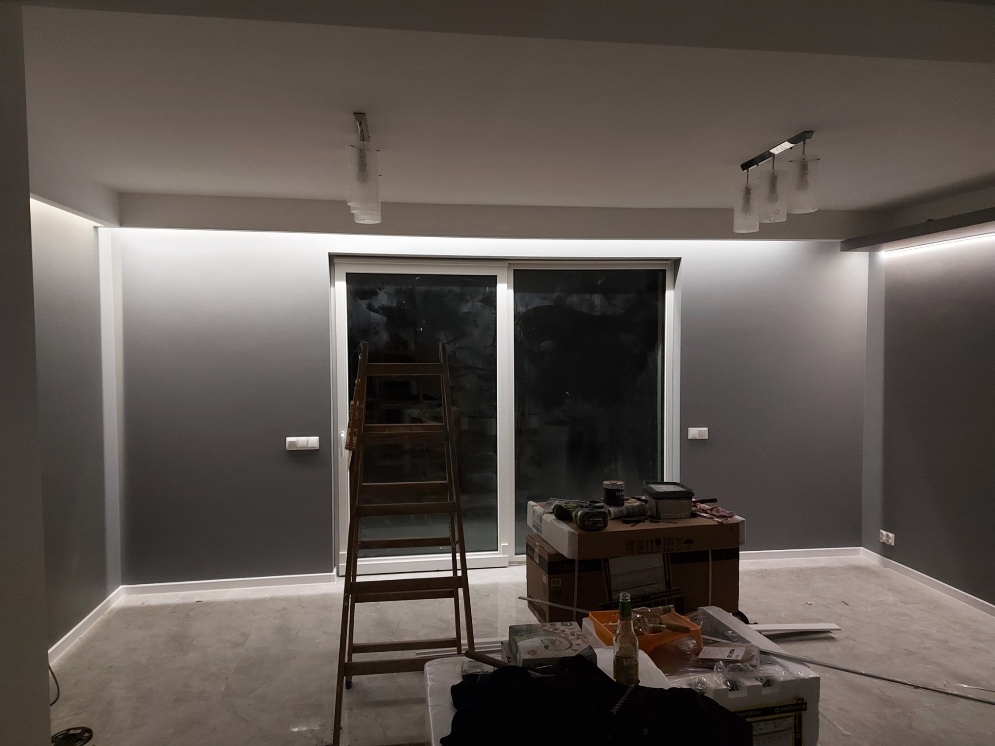 Pokój w trakcie remontu: drabina oparta o okno balkonowe, kartony z narzędziami i materiałami budowlanymi, częściowo pomalowane szare ściany z oświetleniem LED pod sufitem.