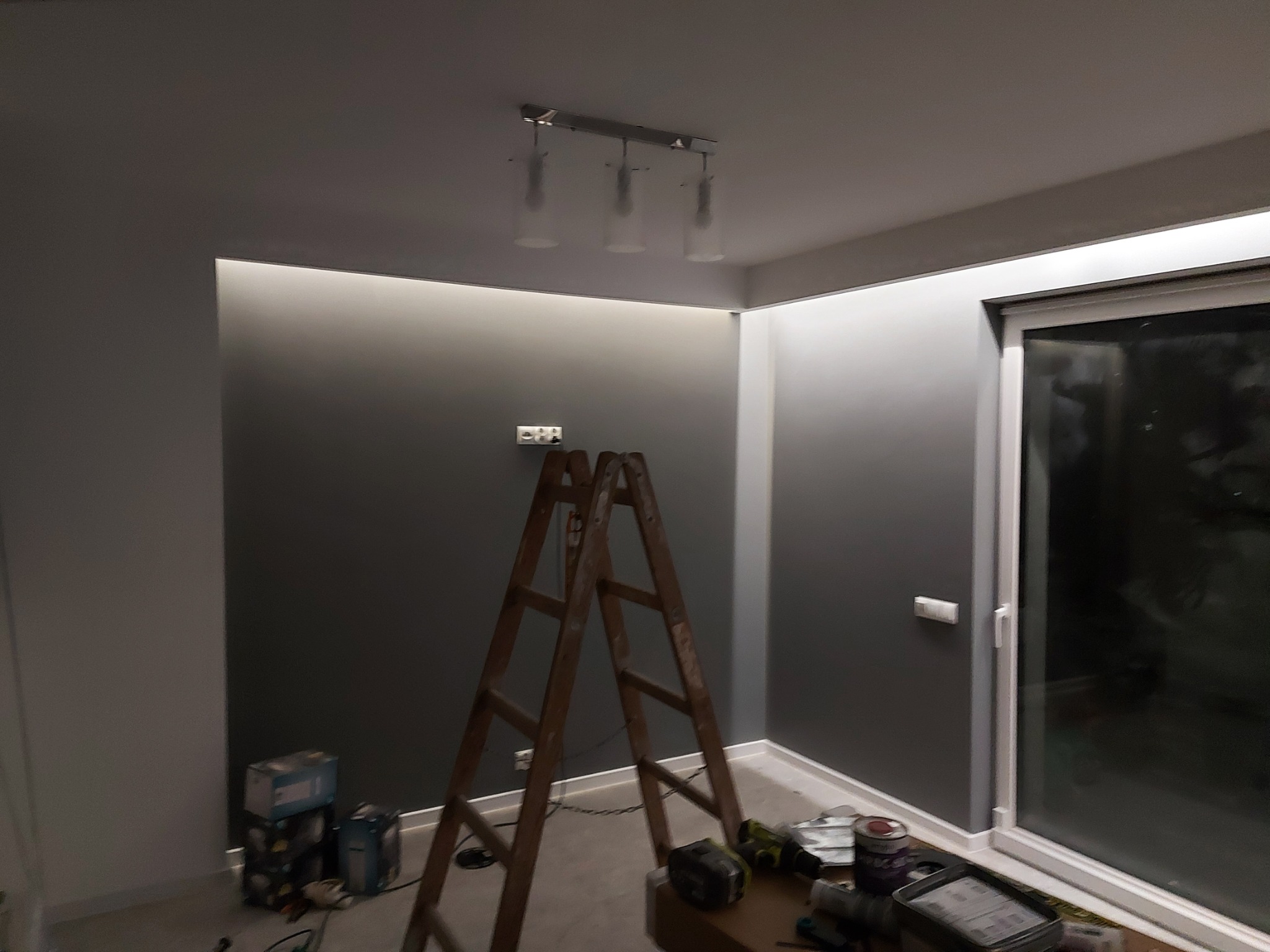 Wnętrze w trakcie remontu: drabina oparta o ścianę, widoczne narzędzia malarskie i puszki z farbą, oświetlenie sufitowe z trzema lampami, szare ściany z listwami LED.