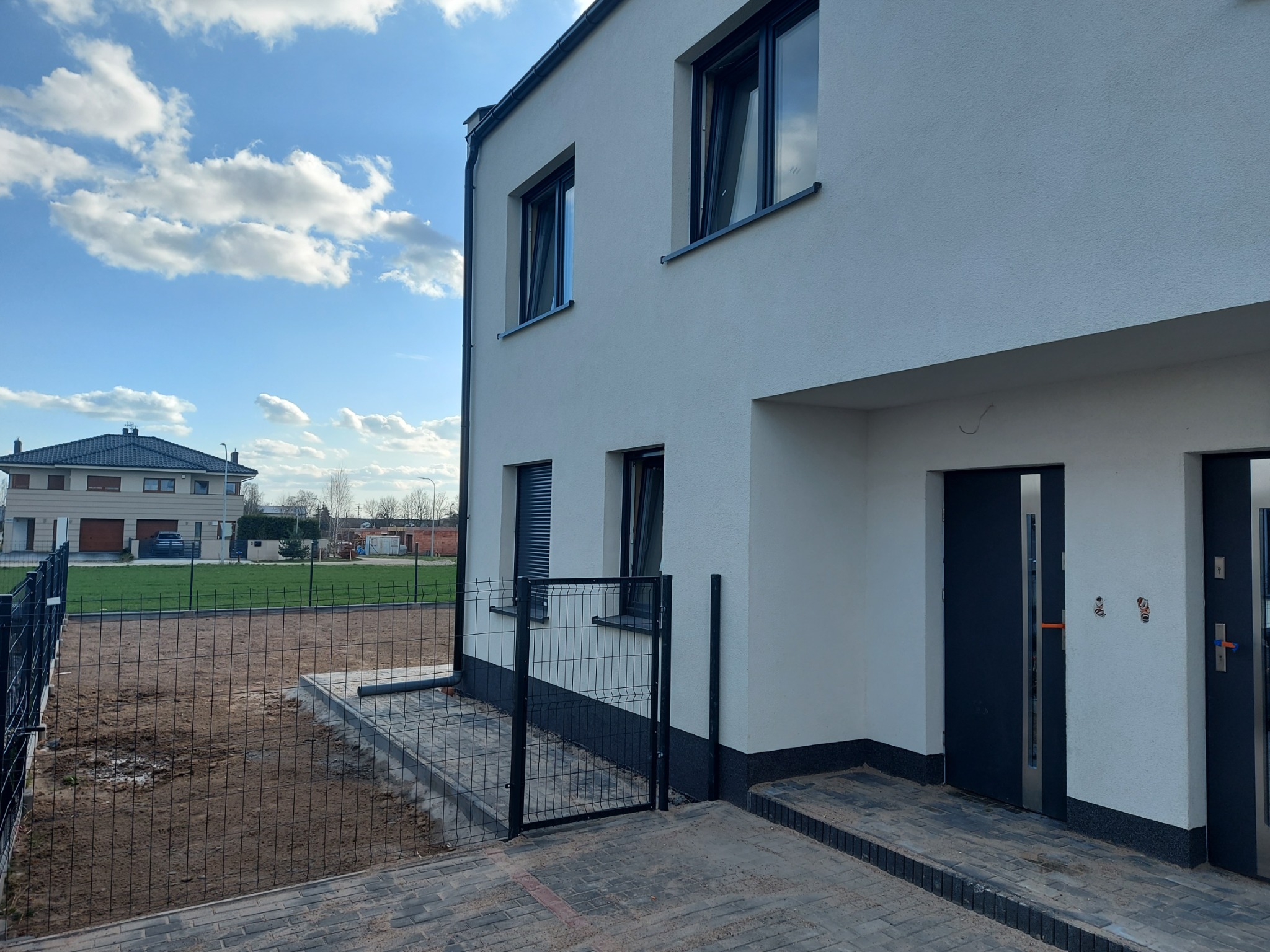 Nowo wybudowany dom jednorodzinny z białym tynkiem, czarnymi oknami i drzwiami wejściowymi, widoczny fragment ogrodzenia panelowego i sąsiedni dom w tle, błękitne niebo z chmurami.