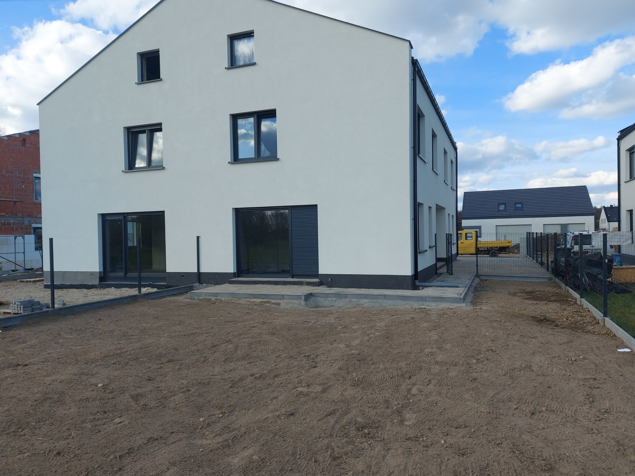 Nowoczesny, dwukondygnacyjny dom z białymi ścianami i dużymi oknami, widoczny z perspektywy przygotowywanego terenu pod trawnik, z żółtą ciężarówką w tle.