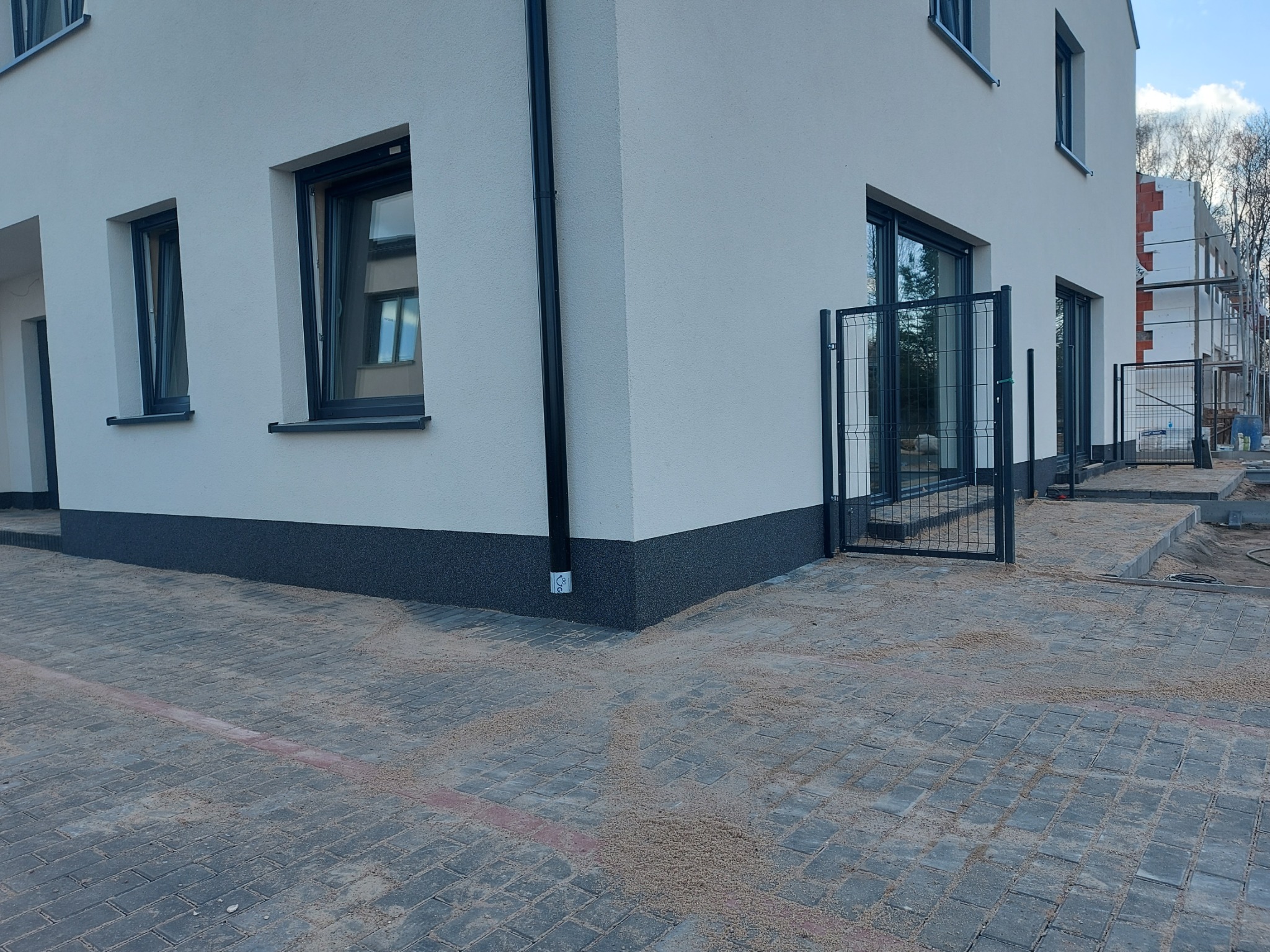 Nowoczesny, biały dom jednorodzinny z szarą podmurówką, czarnymi ramami okiennymi i grafitową, metalową bramką wejściową, widok z zewnątrz na kostkę brukową z piaskiem. W tle widać kolejny budynek...