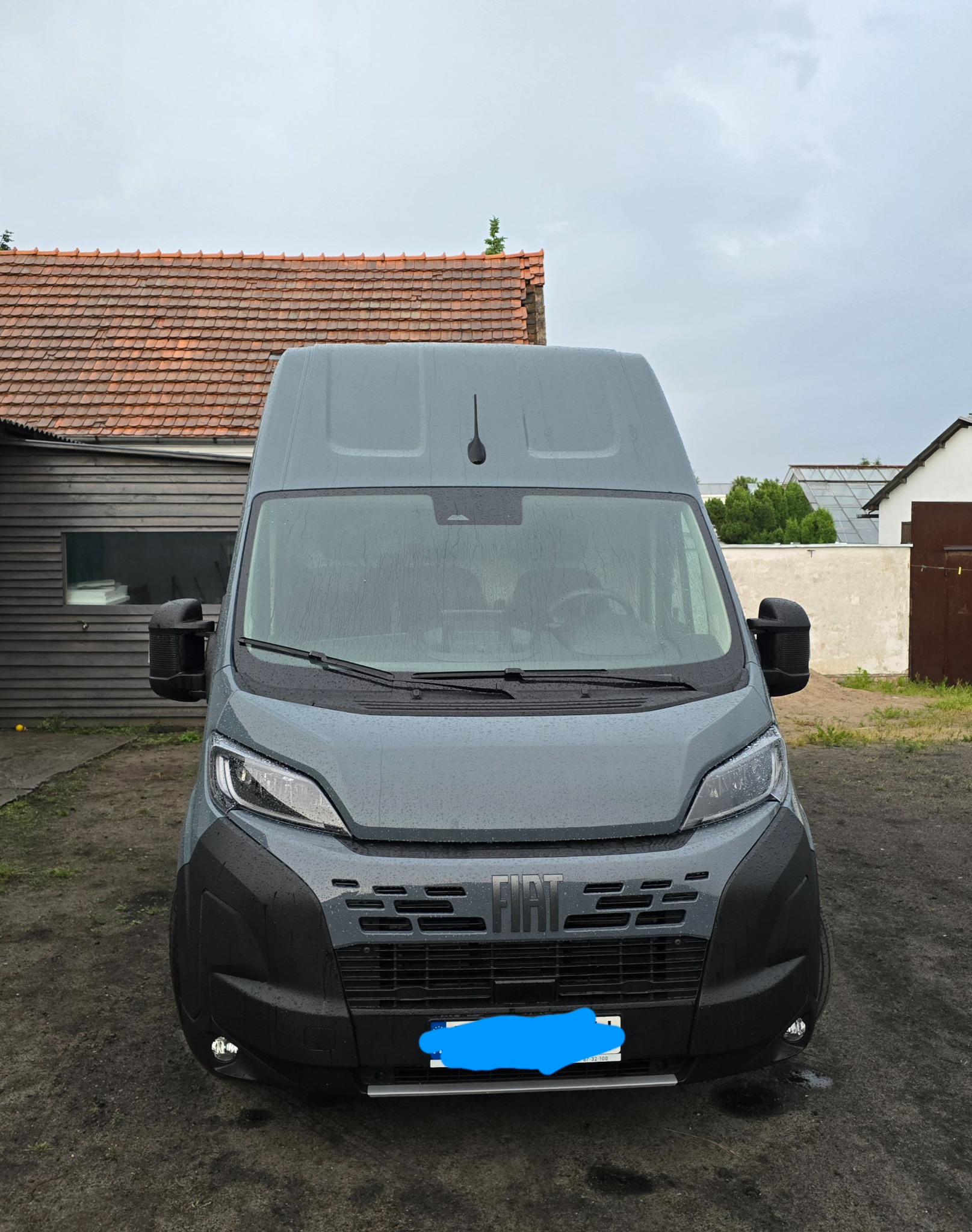 Szary bus marki Fiat stoi na zewnątrz w pochmurny dzień. Widoczne krople deszczu na karoserii. W tle budynek z czerwoną dachówką i fragment białego muru.