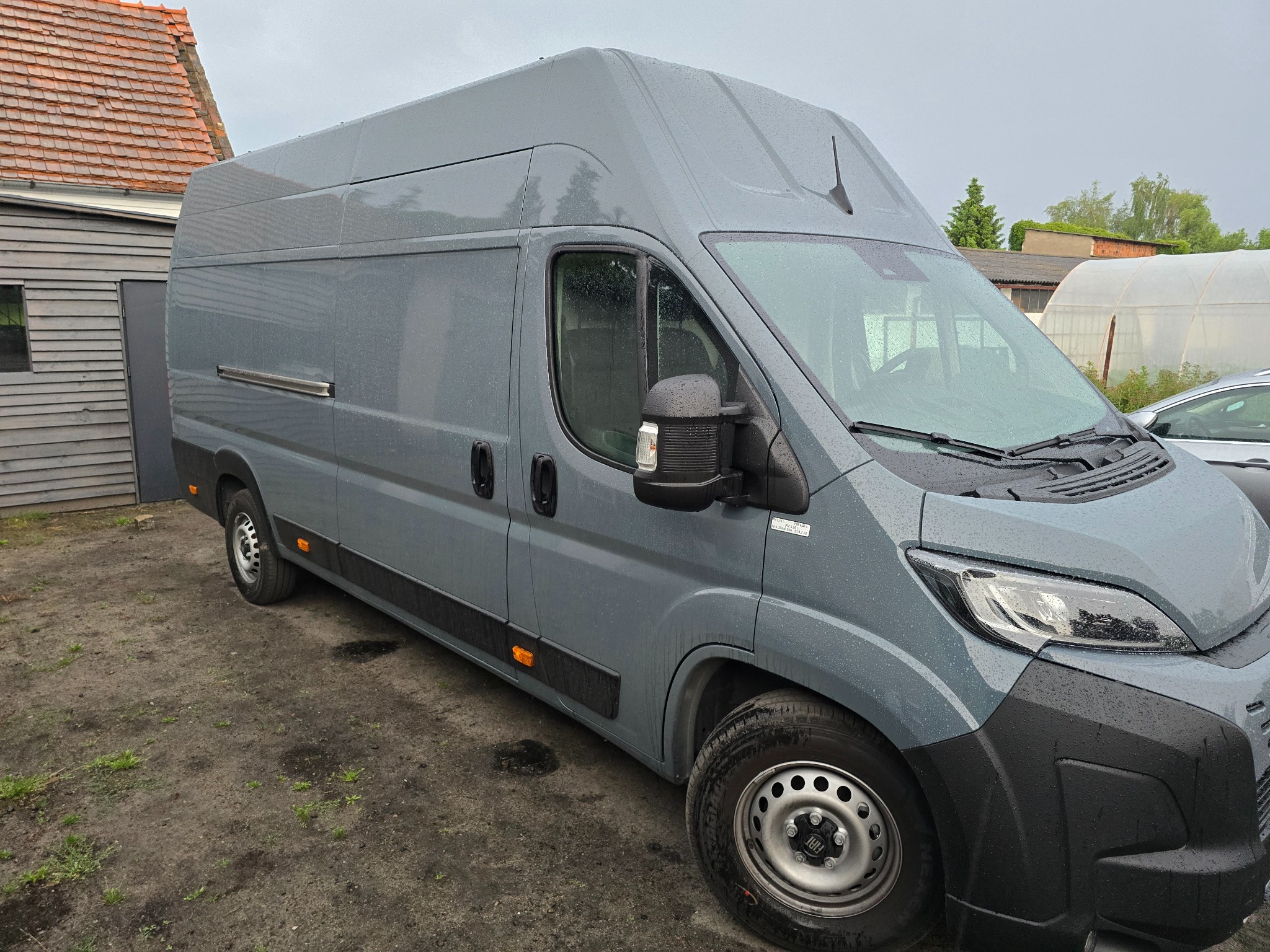 Szary bus dostawczy Fiat Ducato na zewnątrz w deszczowy dzień. Widoczne detale: wysoki dach, relingi boczne, czarne listwy ochronne. Tło: budynek gospodarczy i fragment dachu.