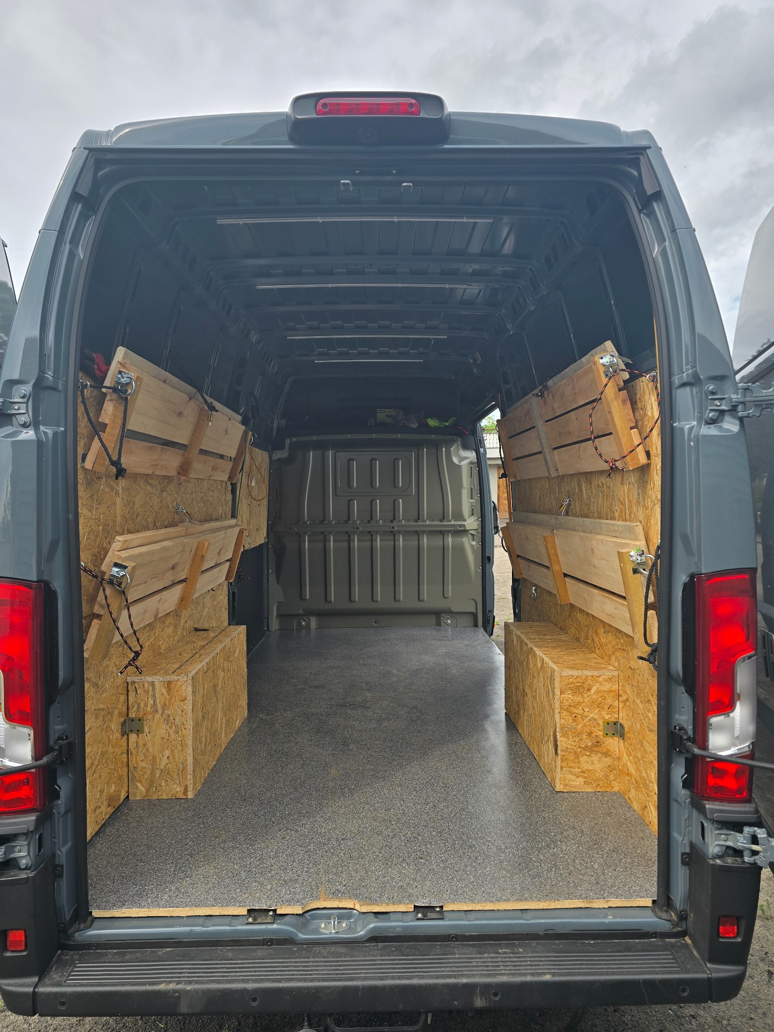 Wnętrze szarego busa z zabudową z płyty OSB: półki i skrzynie po bokach, zabezpieczone linami. Widok z tyłu pojazdu, światło dzienne, szara podłoga.