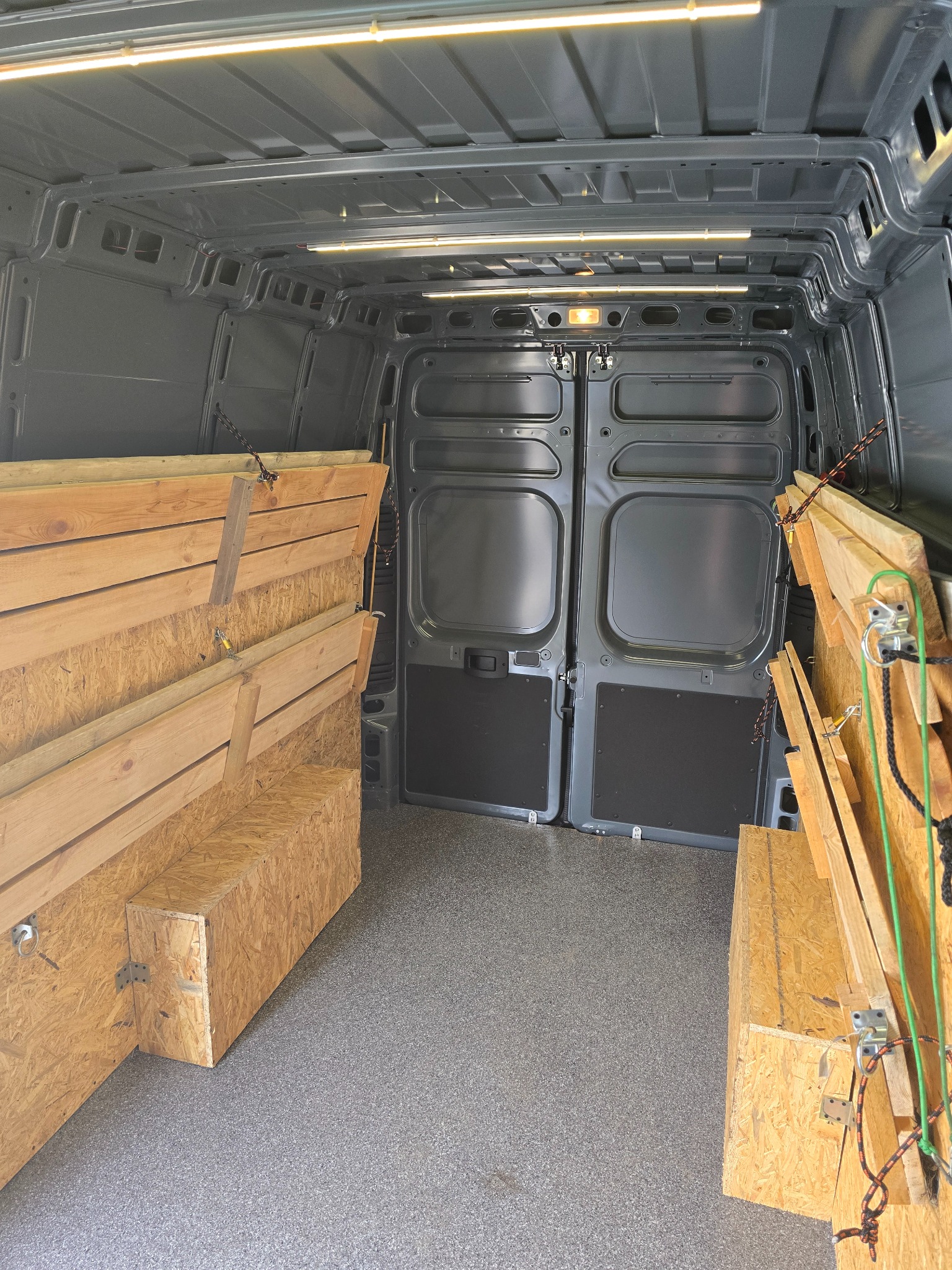Wnętrze szarego busa z zamontowanymi, drewnianymi półkami i zabezpieczeniami ładunku po bokach, gotowe do transportu. Oświetlenie sufitowe.