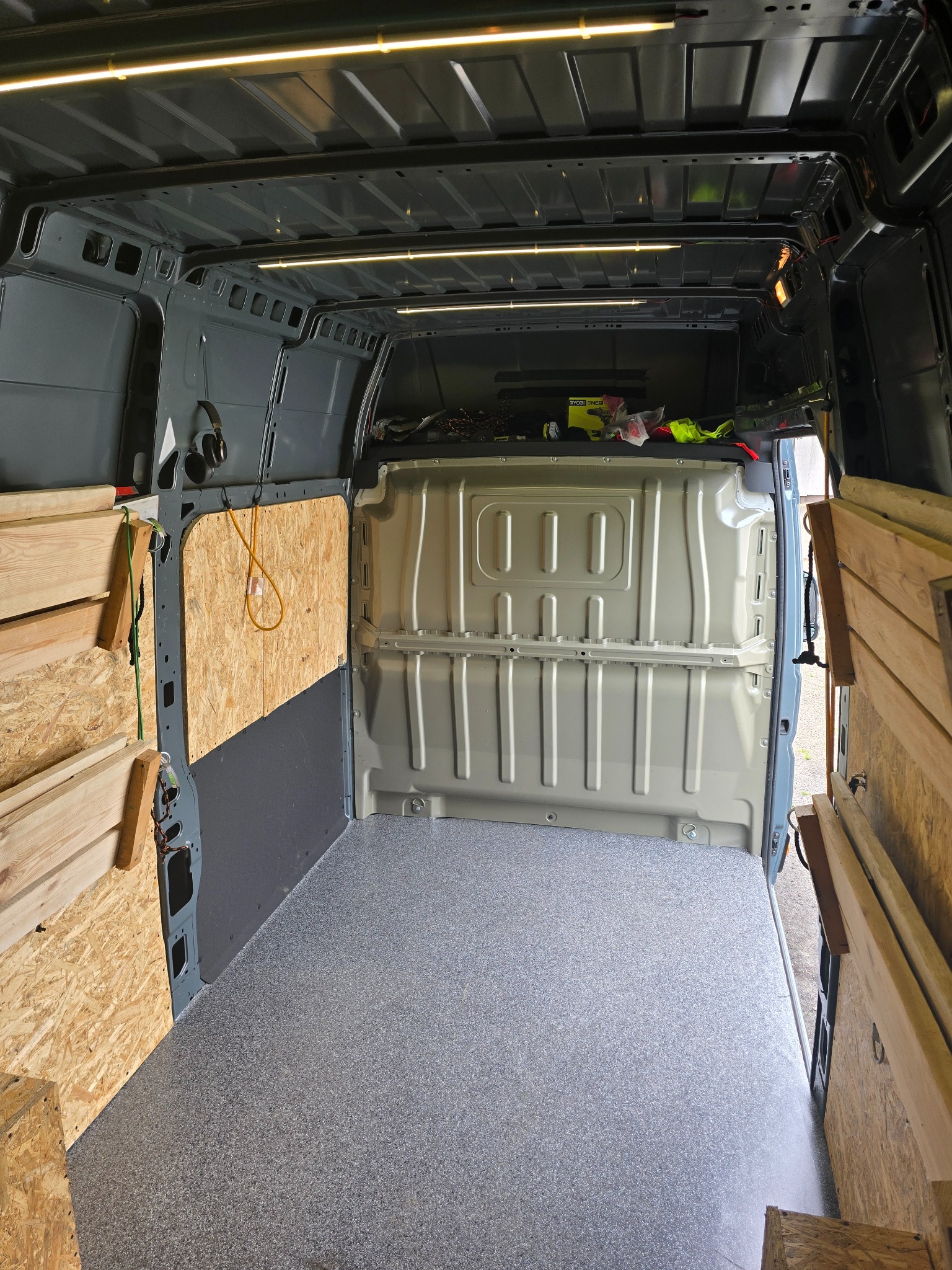 Wnętrze szarego busa z zamontowanymi panelami z płyty OSB na bokach, oświetleniem LED i szarą podłogą. Widoczne narzędzia i akcesoria w tylnej części pojazdu.