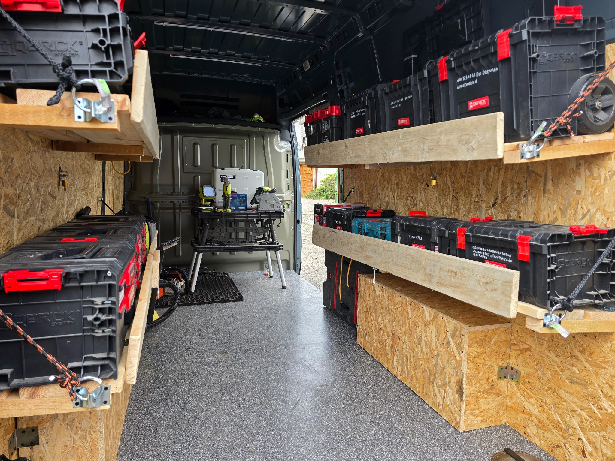 Wnętrze busa z drewnianymi półkami, na których umieszczono skrzynki narzędziowe marki Qbrick. Widoczne narzędzia na stole warsztatowym. Organizacja przestrzeni roboczej.
