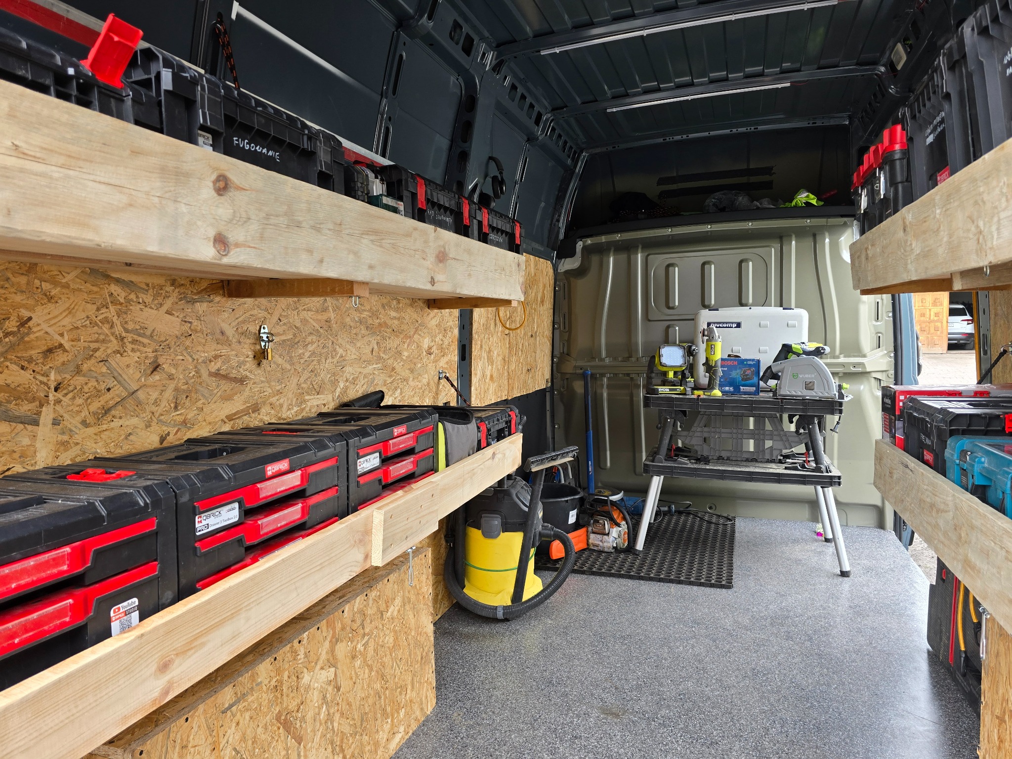 Wnętrze busa z zamontowanymi drewnianymi półkami i skrzynkami narzędziowymi, z widocznym sprzętem na stole warsztatowym. Organizacja przestrzeni roboczej.