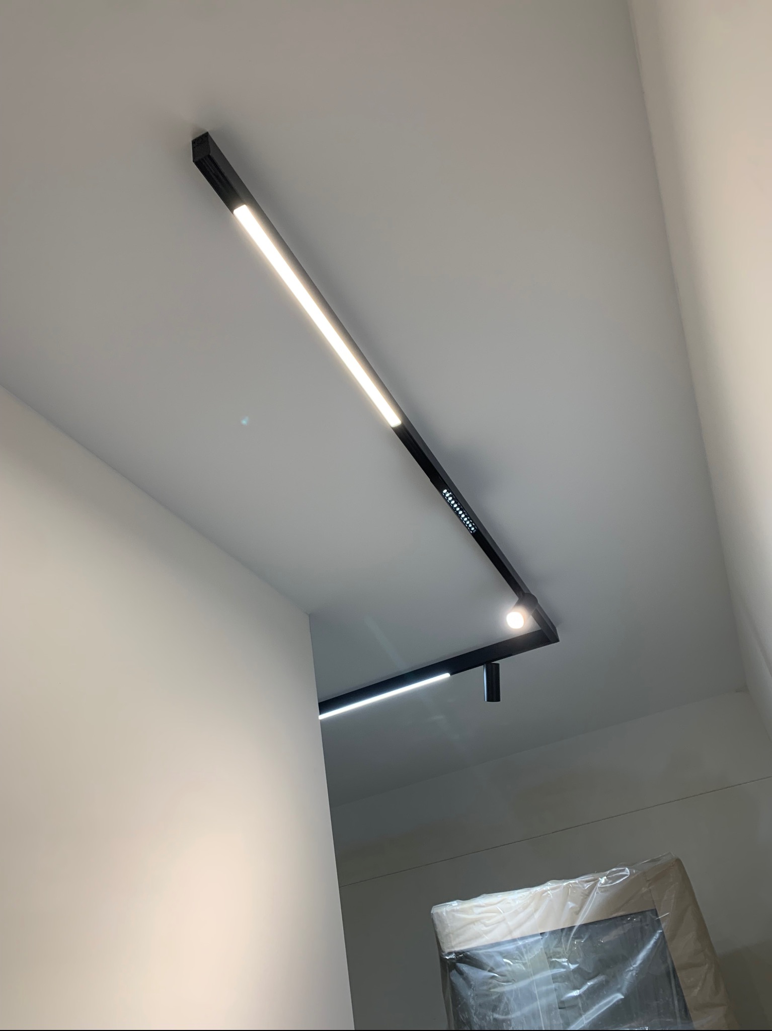 Nowoczesne oświetlenie szynowe w kolorze czarnym, zamontowane na białym suficie, z widocznymi diodami LED i punktowym reflektorem. W tle ściana i zabezpieczony folią mebel.