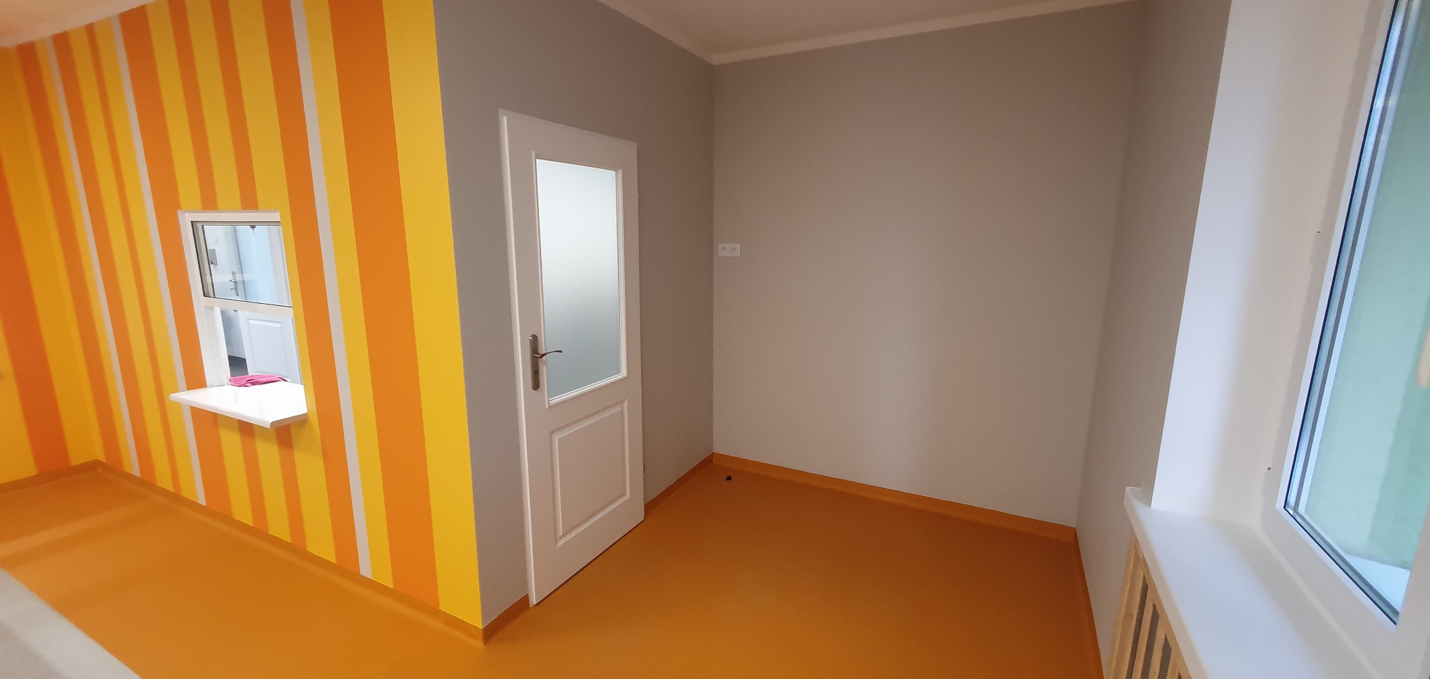 Pomalowane wnętrze pokoju z pomarańczowo-żółtymi pasami na jednej ścianie, szarą ścianą z białą futryną drzwi i pomarańczową podłogą z żywicy.