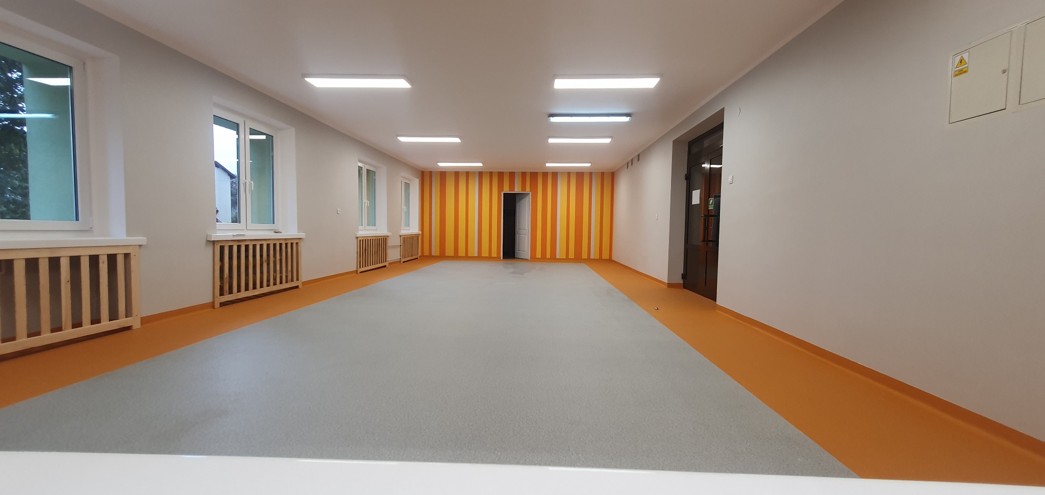 Wnętrze odnowionego korytarza z szarą posadzką, pomarańczową listwą przypodłogową i ścianą w pomarańczowo-żółte pasy, widoczne okna z drewnianymi osłonami grzejników.