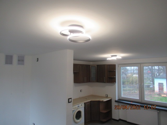 Wnętrze mieszkania w trakcie remontu: widok na sufit z nowoczesnym oświetleniem LED, fragment ściany z kratkami wentylacyjnymi, zabudowa kuchenna w ciemnym odcieniu drewna oraz pralka w rogu...