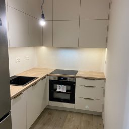 Jest Poziom - Nowoczesna, narożna kuchnia z jasnymi, matowymi frontami szafek, drewnianym blatem, czarnym zlewem i piekarnikiem; wisząca żarówka bez klosza.