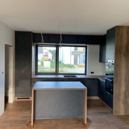 Jest Poziom - Nowoczesna kuchnia z wyspą w trakcie wykańczania, widoczne niedokończone ściany i wystające kable, dominują ciemne kolory i drewniane akcenty.