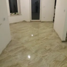 Ułożona podłoga z płytek ceramicznych w odcieniach beżu i brązu, widoczne fugi, białe ściany w trakcie wykańczania, okno i drzwi balkonowe z widoczną sylwetką osoby.