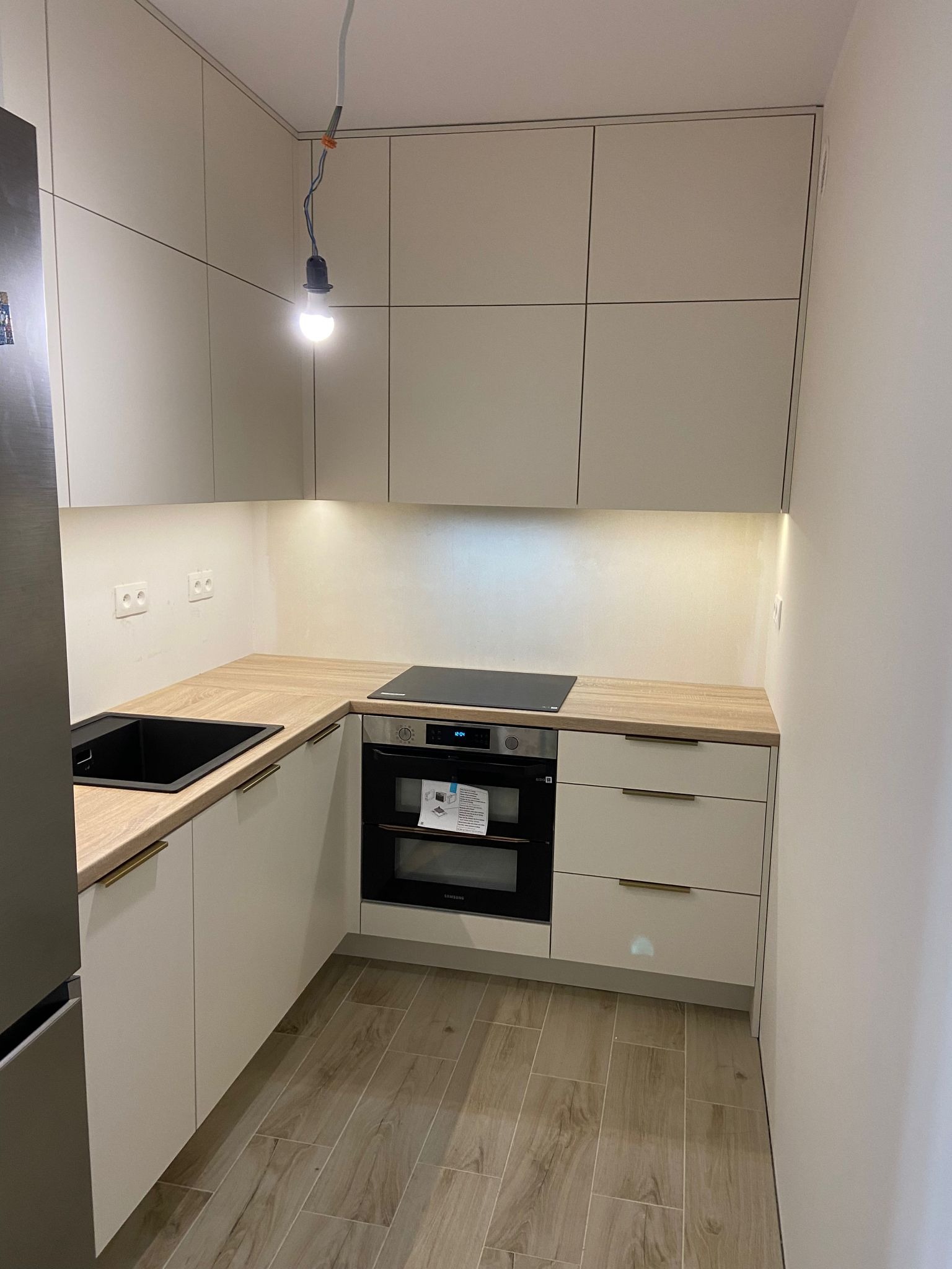 Nowoczesna, narożna kuchnia z jasnymi, matowymi frontami szafek, drewnianym blatem, czarnym zlewem i piekarnikiem; wisząca żarówka bez klosza.