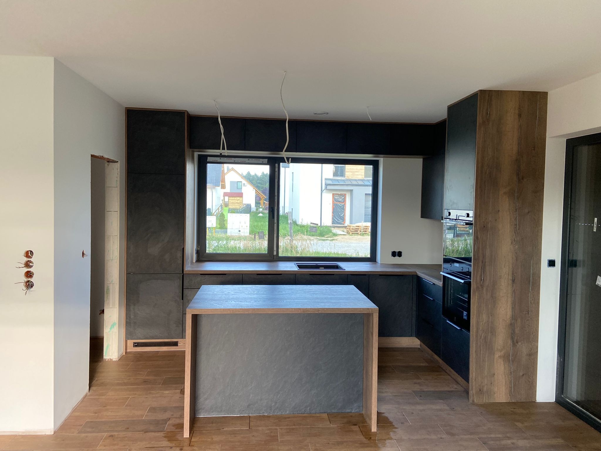 Nowoczesna kuchnia z wyspą w trakcie wykańczania, widoczne niedokończone ściany i wystające kable, dominują ciemne kolory i drewniane akcenty.