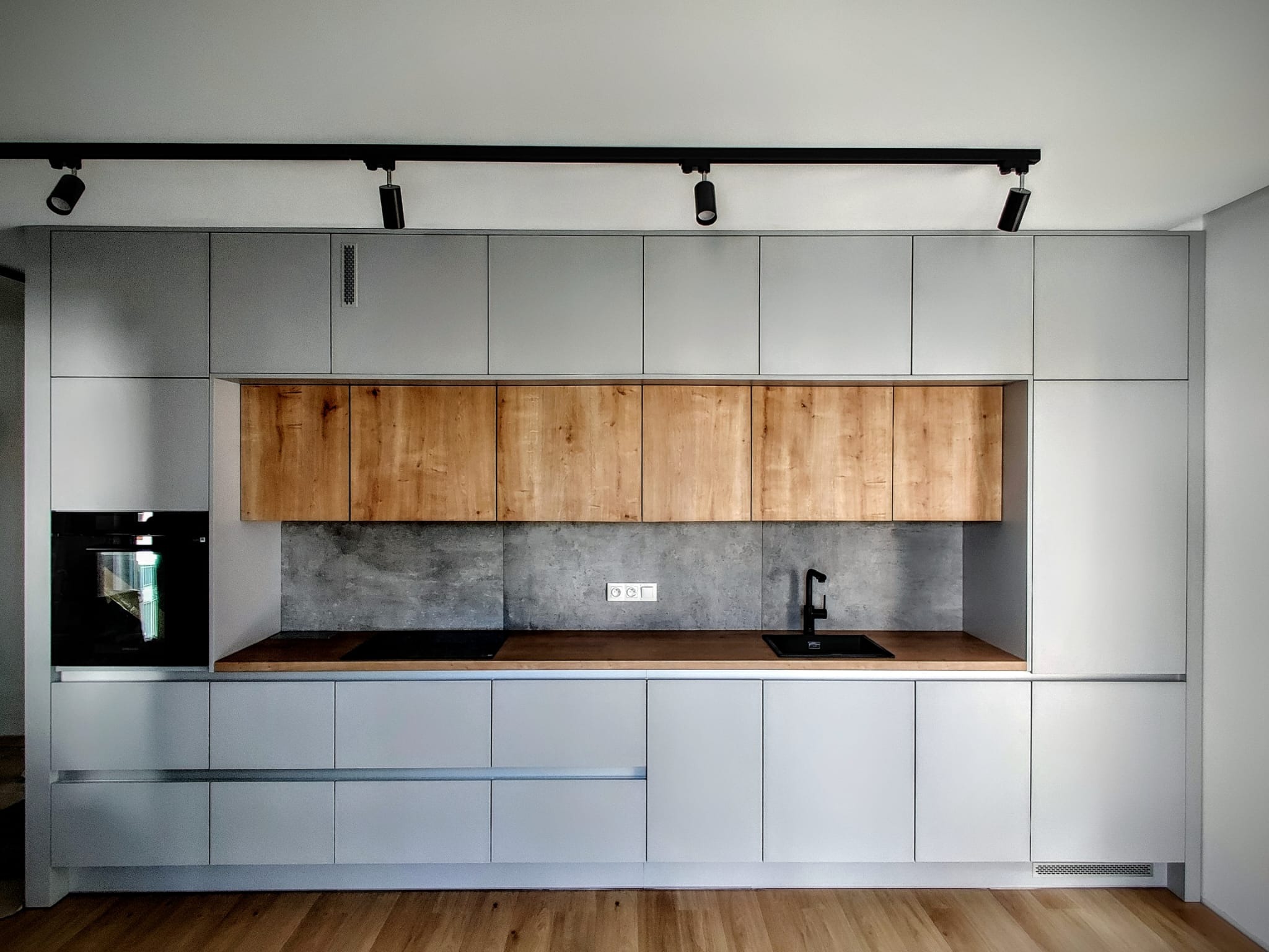 Nowoczesna kuchnia z szarymi szafkami, drewnianymi akcentami i czarną armaturą, oświetlona reflektorami na szynoprzewodzie.