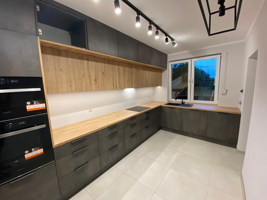 Nowoczesna kuchnia w odcieniach szarości z drewnianymi akcentami, oświetleniem punktowym i dużym oknem z widokiem na zieleń.