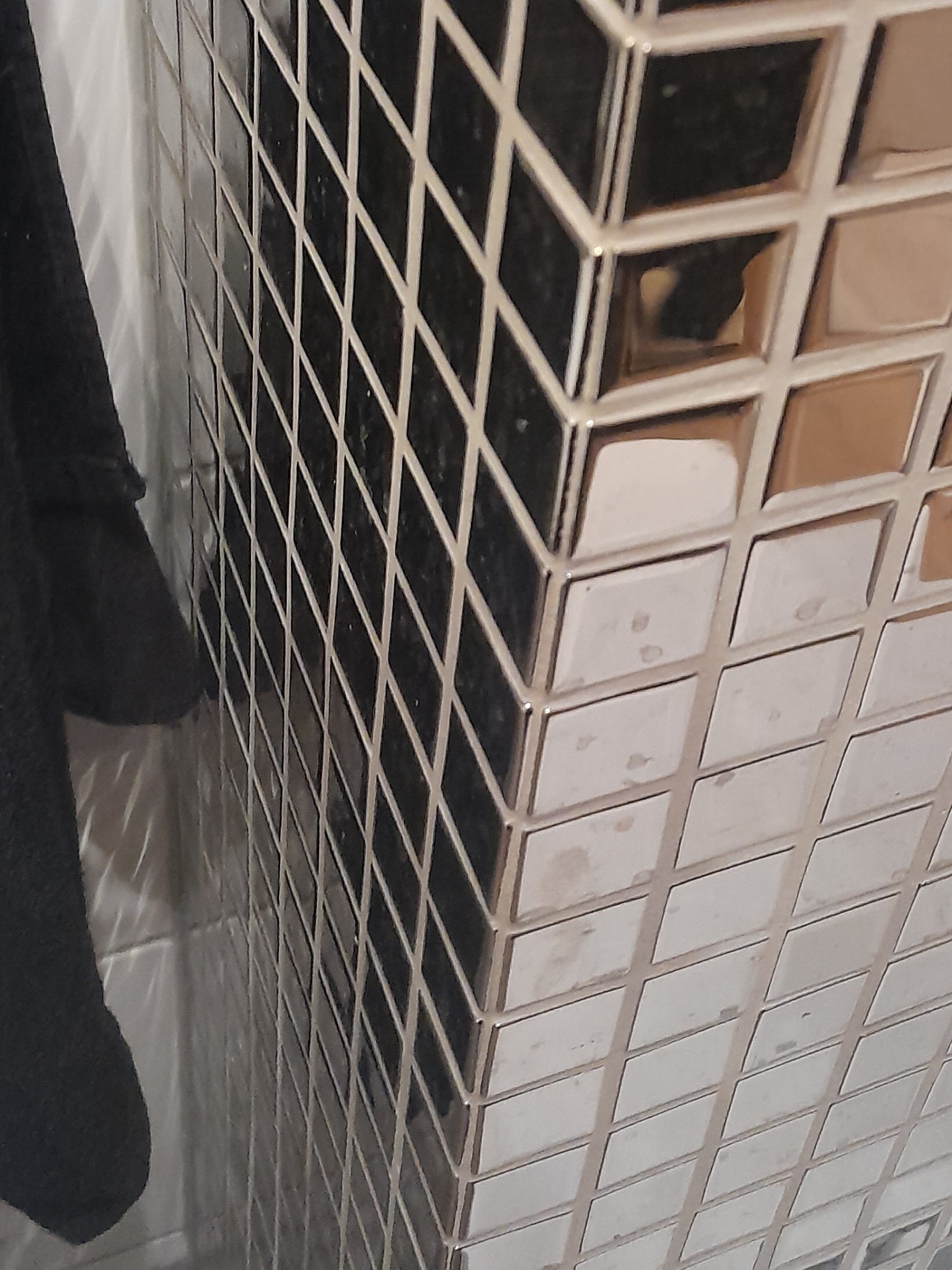 Ściana wykończona mozaiką z małych, lustrzanych płytek w kształcie kwadratów, ułożonych pod kątem, z widocznym fragmentem ciemnego materiału po lewej stronie.