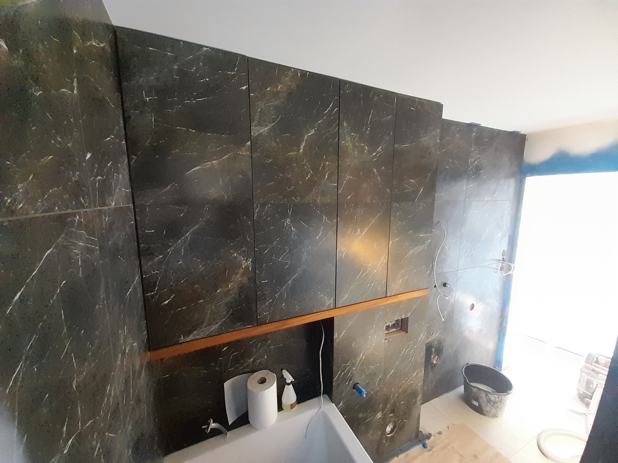 Wnętrze łazienki w trakcie remontu: ściany wyłożone ciemnymi płytkami imitującymi marmur, widoczna wanna, akcesoria łazienkowe i narzędzia budowlane.