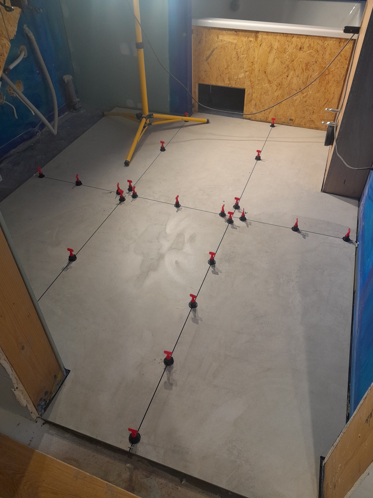 Świeżo wylany, jasnoszary betonowy podkład podłogowy z rozstawionymi, czerwonymi elementami poziomującymi, ustawionymi wzdłuż czarnych sznurków, oświetlony żółtą lampą budowlaną.
