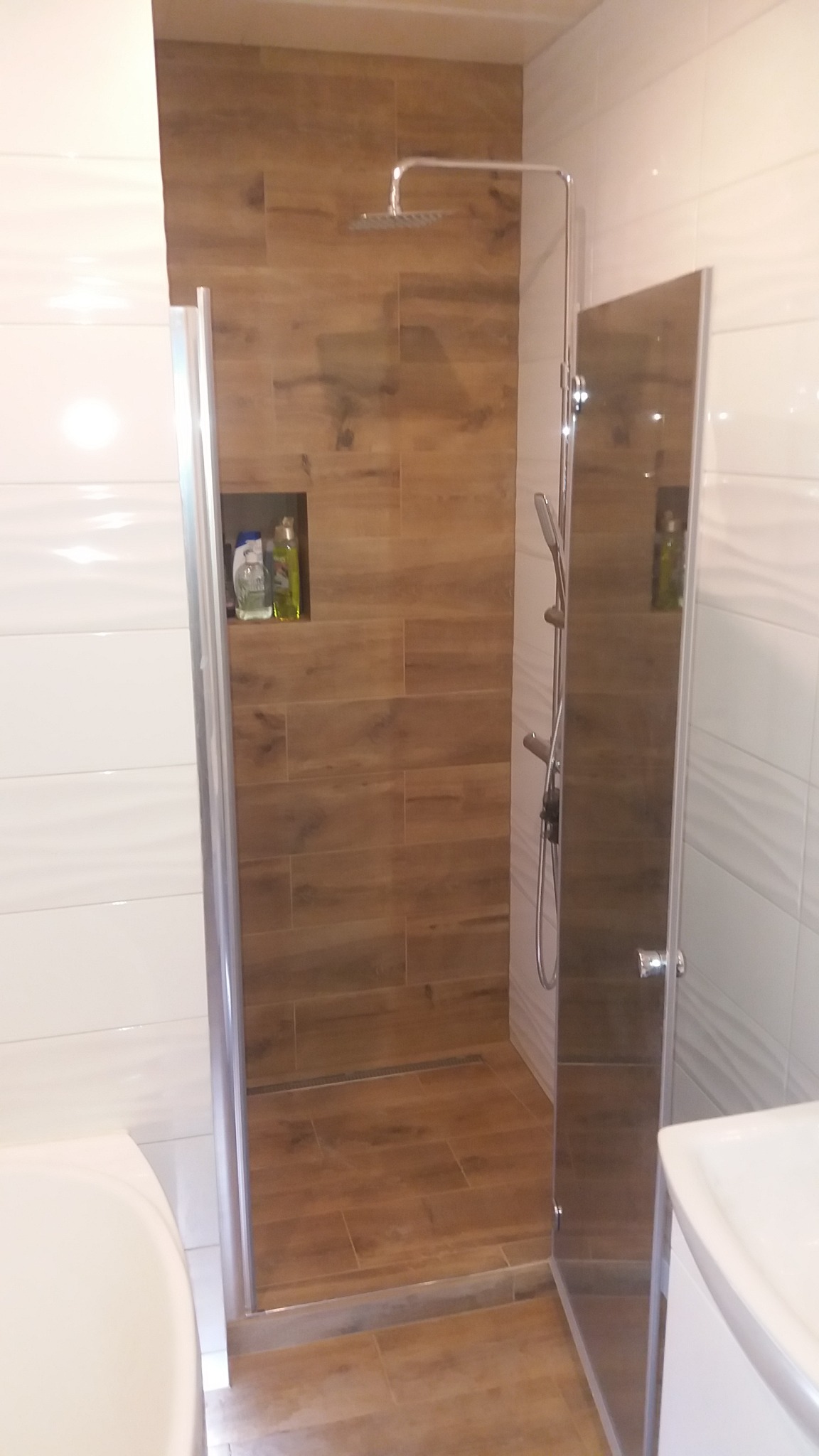 Nowoczesna kabina prysznicowa z brązowymi płytkami imitującymi drewno, wbudowana półka na kosmetyki, chromowana armatura, częściowo otwarta szklana ściana.