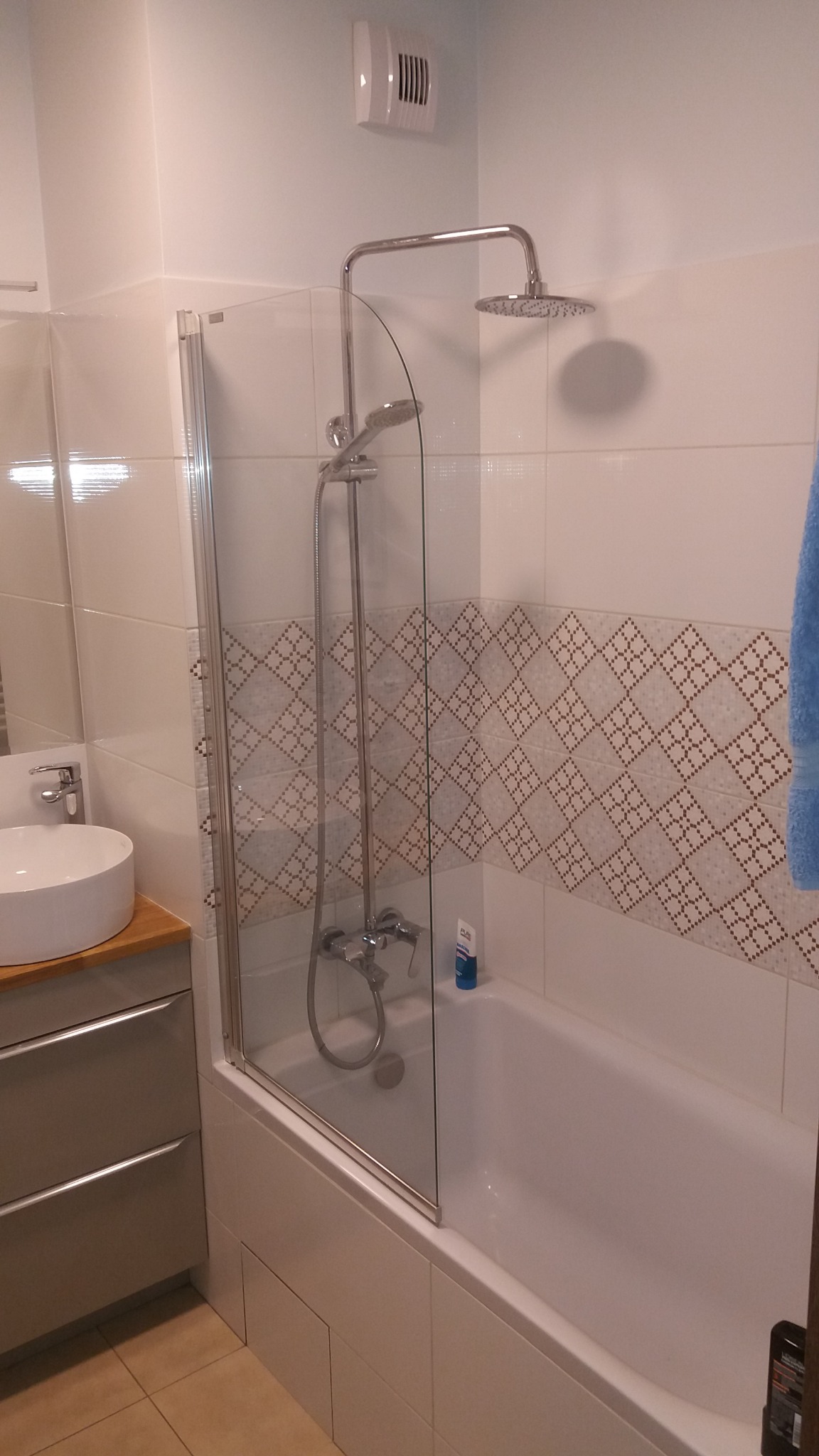 Nowoczesna łazienka z wanną obudowaną białymi płytkami, szklanym parawanem nawannowym, baterią prysznicową i deszczownicą, umywalką nablatową na szafce z szufladami oraz dekoracyjnym pasem płytek...