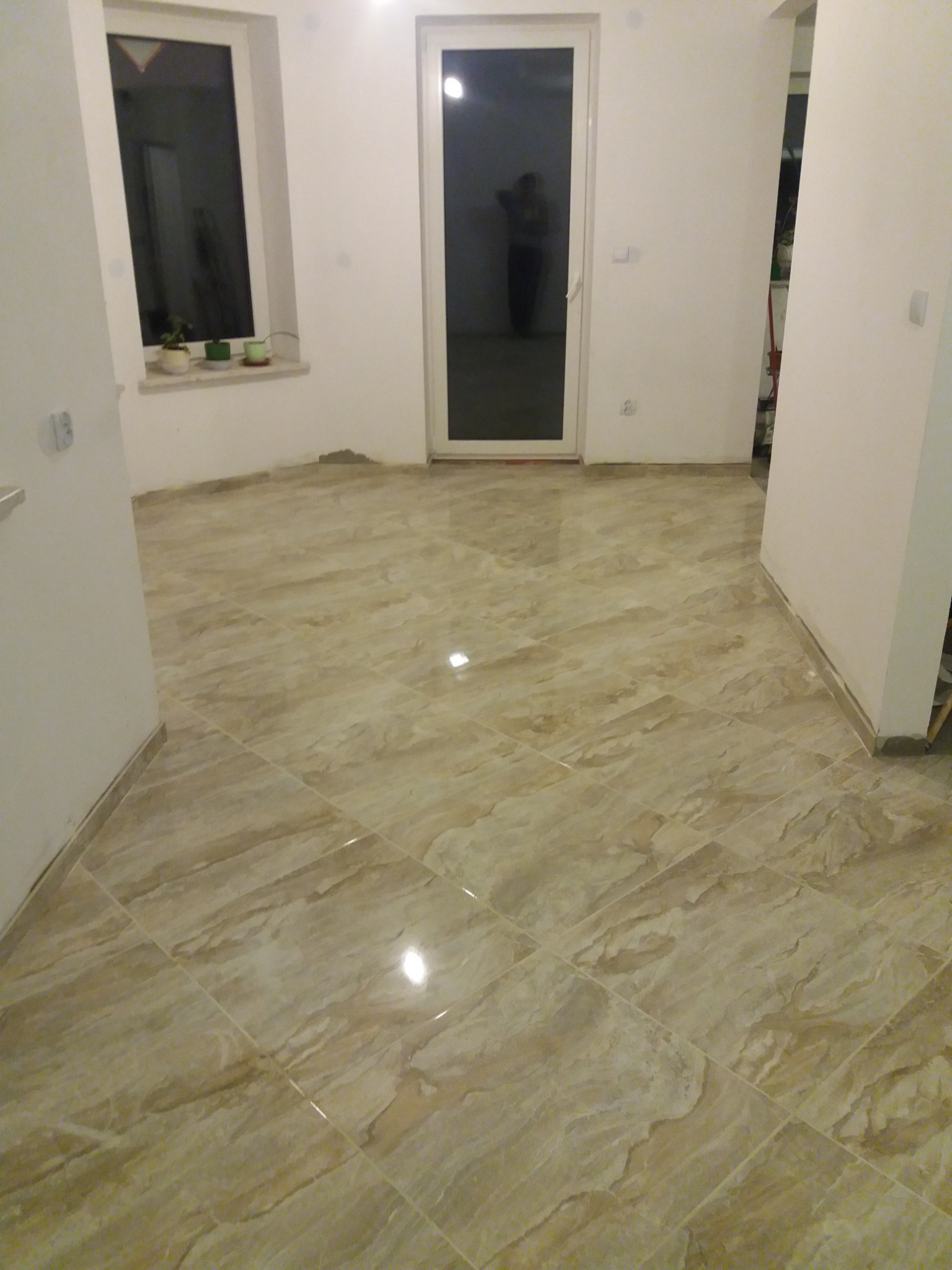 Ułożona podłoga z płytek ceramicznych w odcieniach beżu i brązu, widoczne fugi, białe ściany w trakcie wykańczania, okno i drzwi balkonowe z widoczną sylwetką osoby.