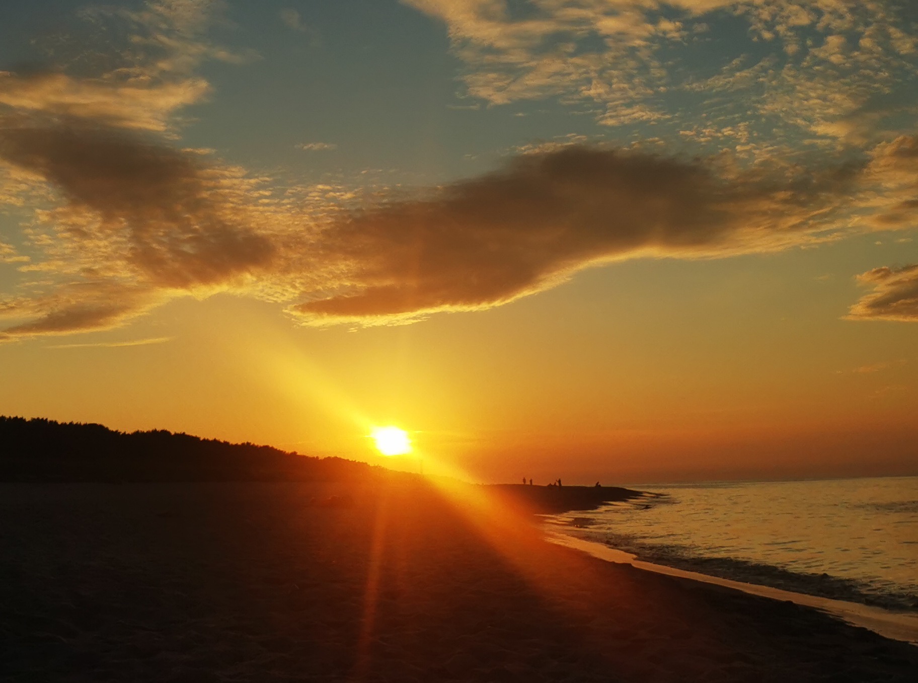 Zachód słońca nad morzem: złote promienie oświetlają plażę, sylwetki ludzi na horyzoncie, malownicze obłoki na niebie. Ciepła kolorystyka.