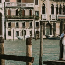 Bierzemy Ślub - Sesje & Reportaże Ślubne - Para młoda w objęciach na drewnianym pomoście w Wenecji, panna młoda w koronkowej sukni z odkrytymi plecami i wiankiem, w tle weneckie budynki i kanały.