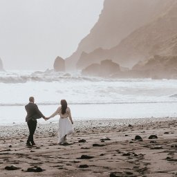 Bierzemy Ślub - Sesje & Reportaże Ślubne - Para młoda trzymająca się za ręce spaceruje po czarnej, kamienistej plaży w Islandii, z widokiem na formacje skalne i wzburzone morze we mgle.