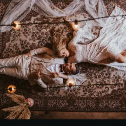 Para młoda leżąca na wzorzystym dywanie, oświetlona ciepłym światłem świec, panna młoda w koronkowej sukni, pan młody w beżowym garniturze, ujęcie z g&oacute;ry.