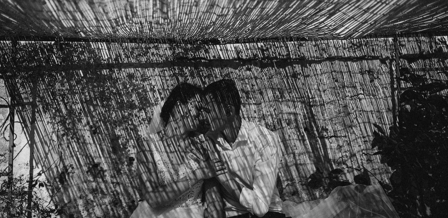 Czarno-białe zdjęcie pary w objęciach, z filtrem z cieni rzucanych przez trzcinową konstrukcję, tworzącym unikalny wzór na ich sylwetkach i tle.