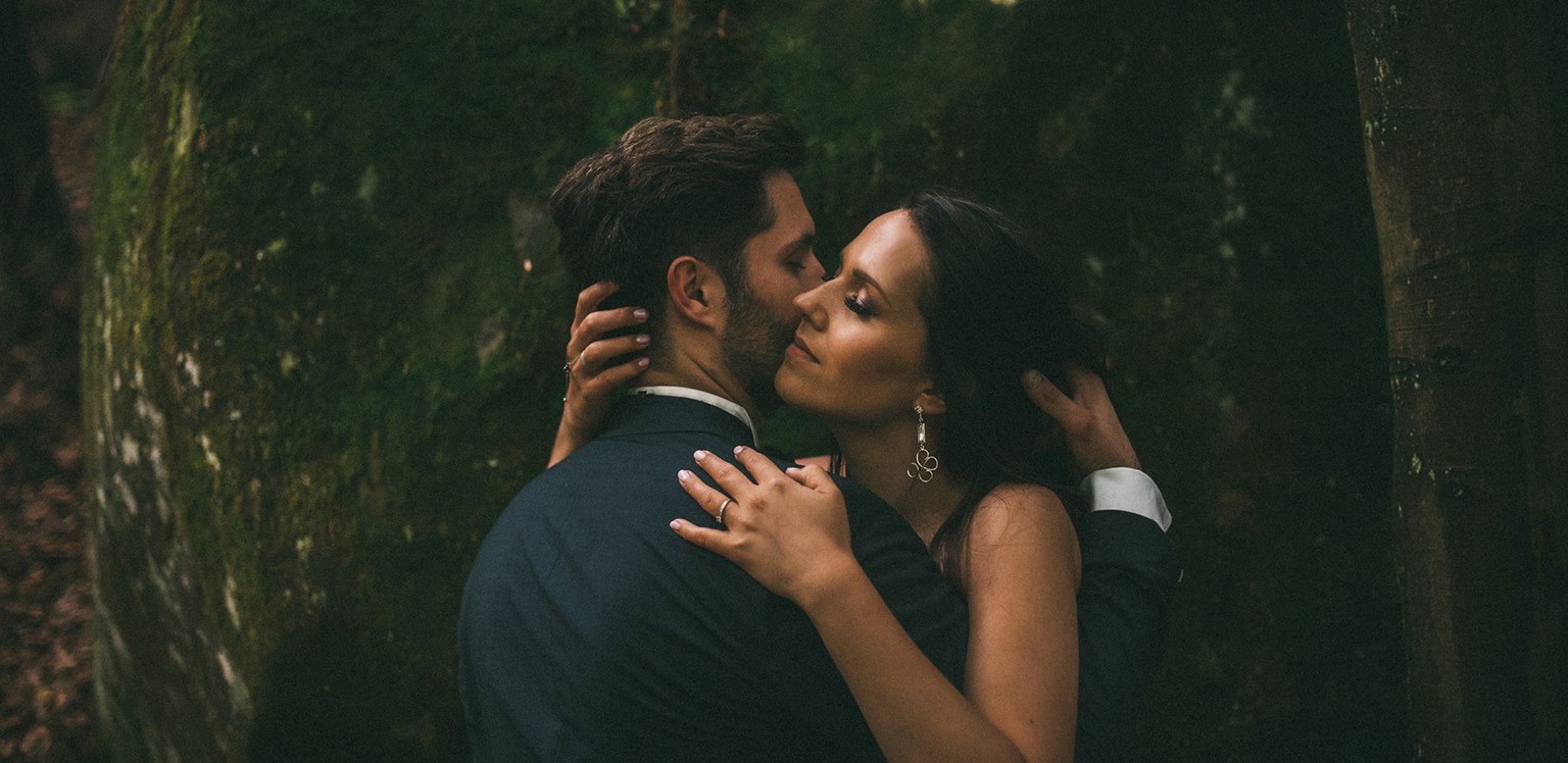 Para narzeczeńska w objęciach na tle omszałego drzewa, ona w sukience z delikatną biżuterią, on w granatowym garniturze.