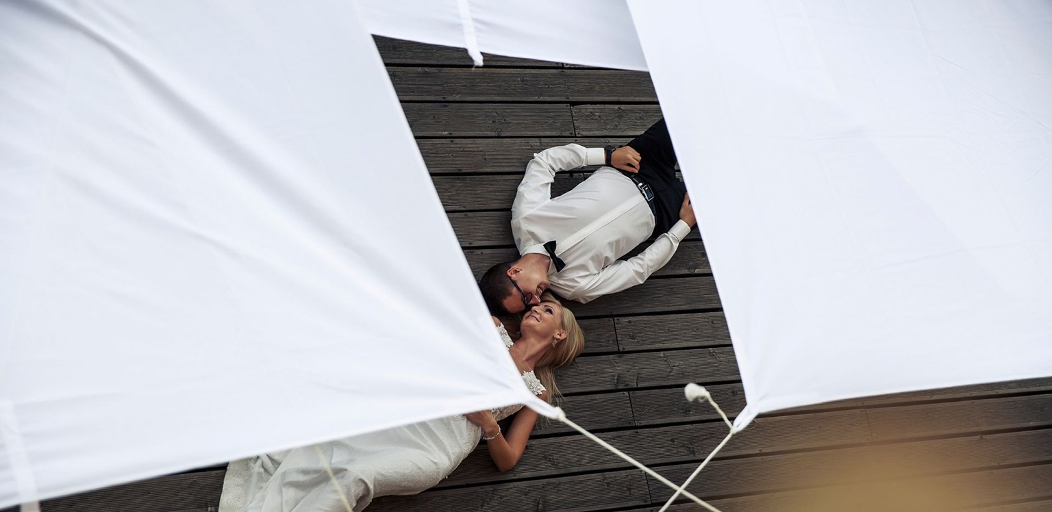 Para młoda leżąca na drewnianym pomoście, widok z góry przesłonięty białymi tkaninami, panna młoda w koronkowej sukni, pan młody w czarnym garniturze z muszką, delikatny uśmiech na twarzach.