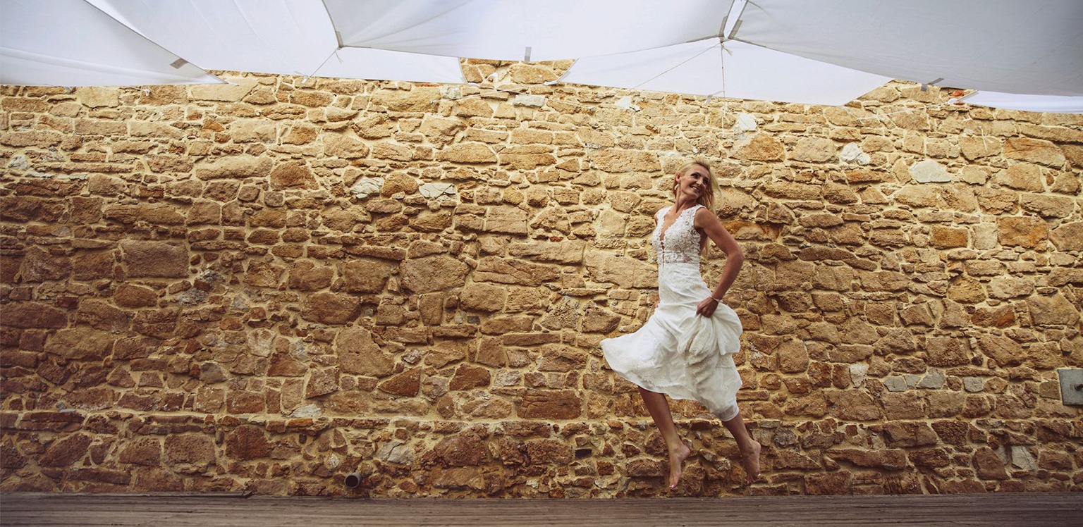 Uśmiechnięta panna młoda w zwiewnej sukni skacze boso na drewnianym podeście, na tle kamiennej ściany i białego baldachimu.