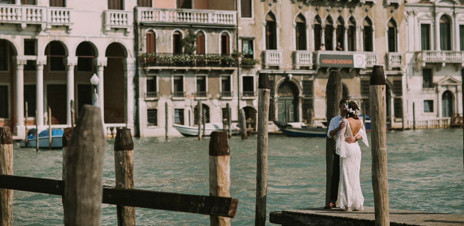 Para młoda w objęciach na drewnianym pomoście w Wenecji, panna młoda w koronkowej sukni z odkrytymi plecami i wiankiem, w tle weneckie budynki i kanały.