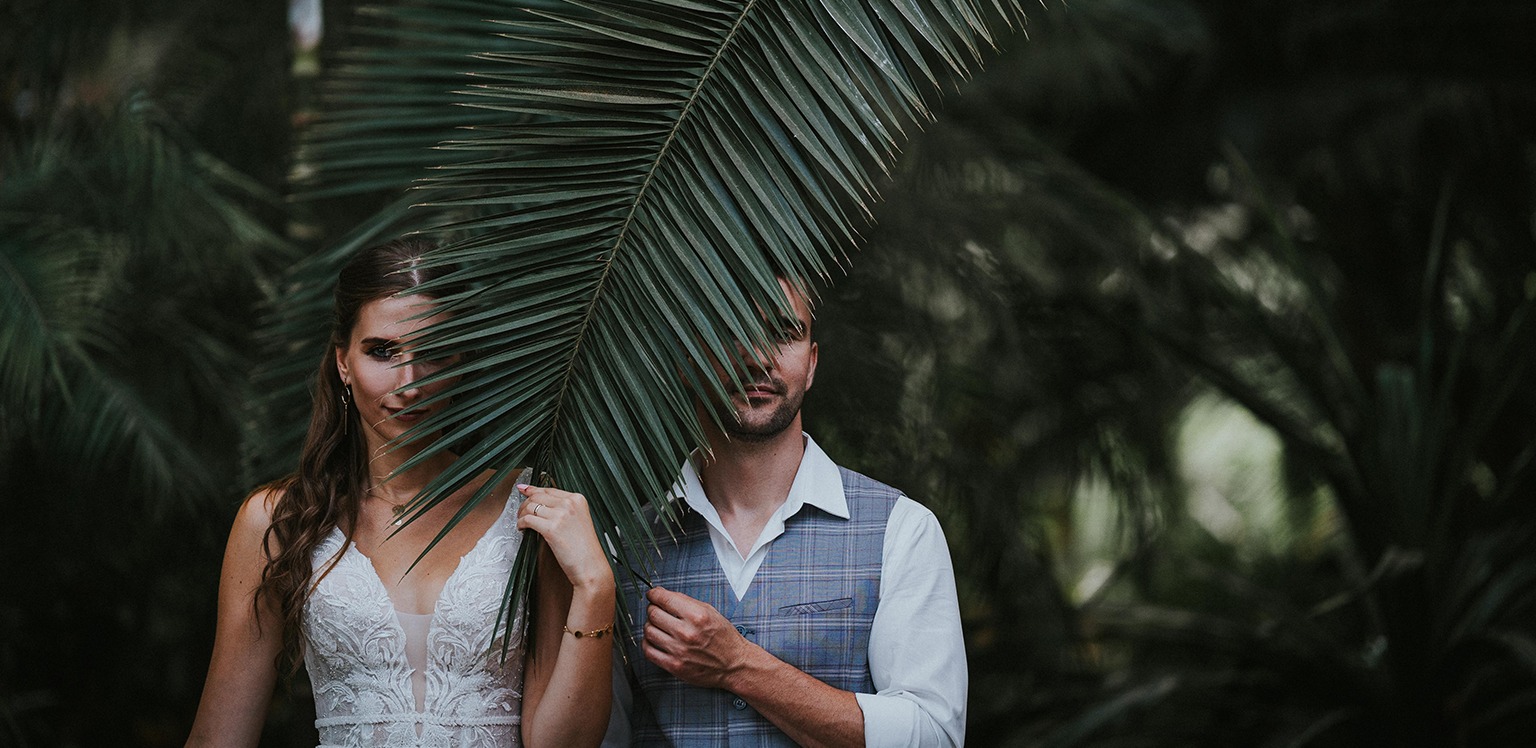 Para młoda częściowo zasłonięta liściem palmy na tle tropikalnej zieleni. Panna młoda w koronkowej sukni, pan młody w kamizelce w kratę.