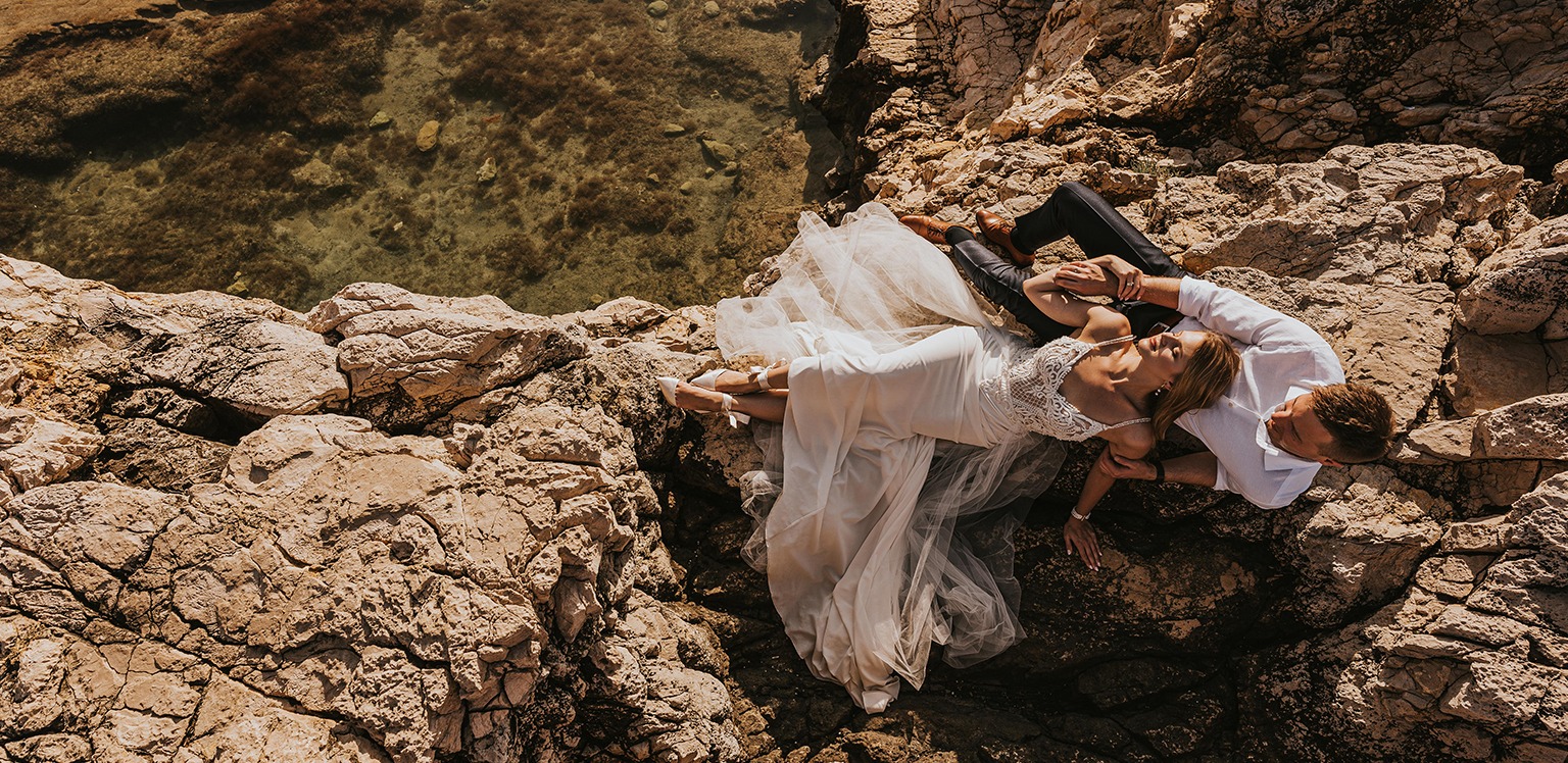 Para w sukni ślubnej i białej koszuli leży na skalistym wybrzeżu w promieniach słońca, z widokiem na czystą wodę.