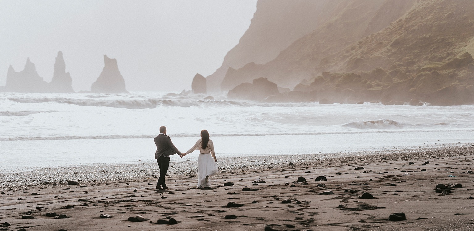 Para młoda trzymająca się za ręce spaceruje po czarnej, kamienistej plaży w Islandii, z widokiem na formacje skalne i wzburzone morze we mgle.