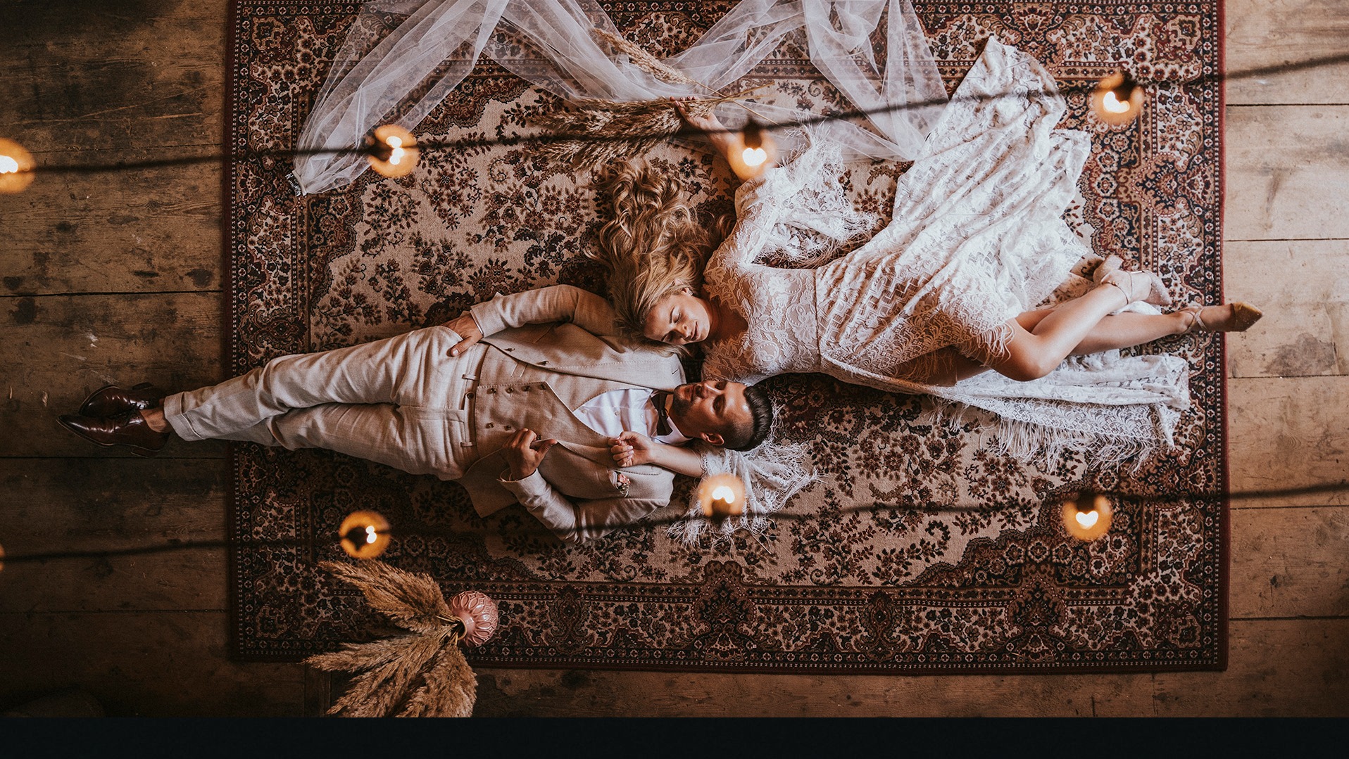 Para młoda leżąca na wzorzystym dywanie, oświetlona ciepłym światłem świec, panna młoda w koronkowej sukni, pan młody w beżowym garniturze, ujęcie z góry.