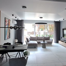 DENKO SPÓŁKA Z OGRANICZONĄ ODPOWIEDZIALNOŚCIĄ - Nowoczesny salon z drewnianą ścianą z lameli, szarą sofą narożną, marmurową podłogą i czarnymi schodami z drewnianymi stopniami.