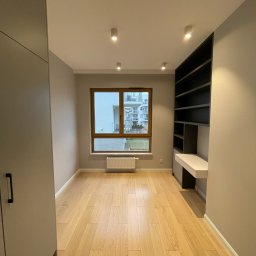 DENKO SPÓŁKA Z OGRANICZONĄ ODPOWIEDZIALNOŚCIĄ - Pokój po remoncie: jasna drewniana podłoga, szara szafa w zabudowie, ciemne regały z biurkiem, okno z widokiem na budynek.