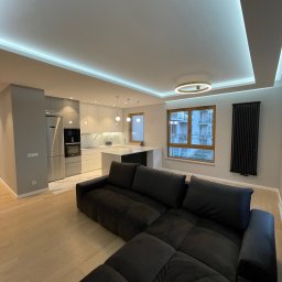 DENKO SPÓŁKA Z OGRANICZONĄ ODPOWIEDZIALNOŚCIĄ - Nowoczesne wnętrze mieszkania z otwartą kuchnią, widok na szarą zabudowę kuchenną z marmurowym blatem i wyspą, czarna sofa na pierwszym planie, oświetlenie LED podsufitowe i okrągła lampa sufitowa,...