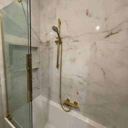 DENKO SPÓŁKA Z OGRANICZONĄ ODPOWIEDZIALNOŚCIĄ - Wykończona łazienka z wanną, marmurowe płytki na ścianach, złota armatura i szklany parawan nawannowy z złotymi detalami.