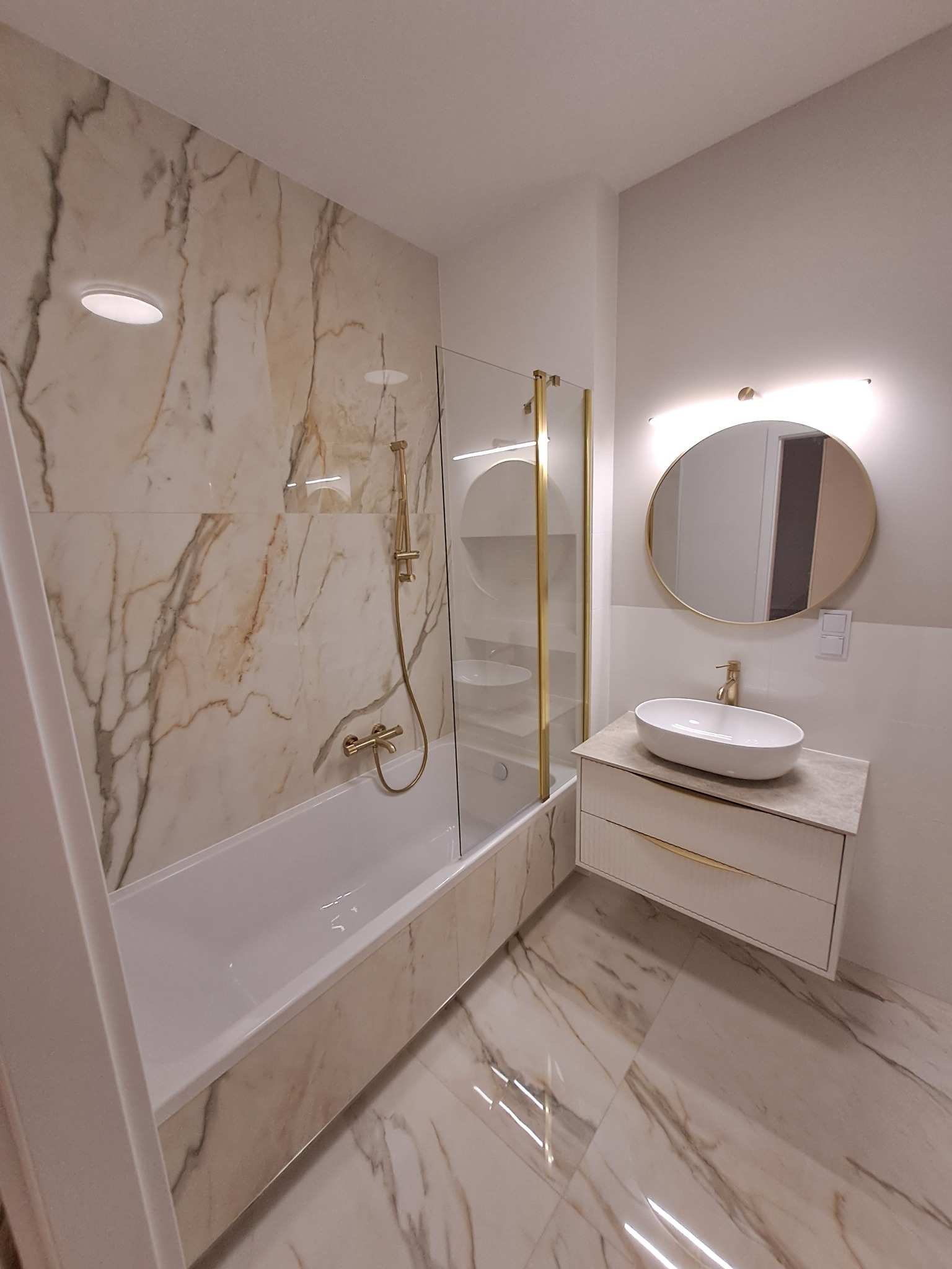 Nowoczesna łazienka z wanną i szklanym parawanem, wykończona płytkami imitującymi marmur, z owalną umywalką na szafce z złotymi akcentami i okrągłym lustrem z podświetleniem.