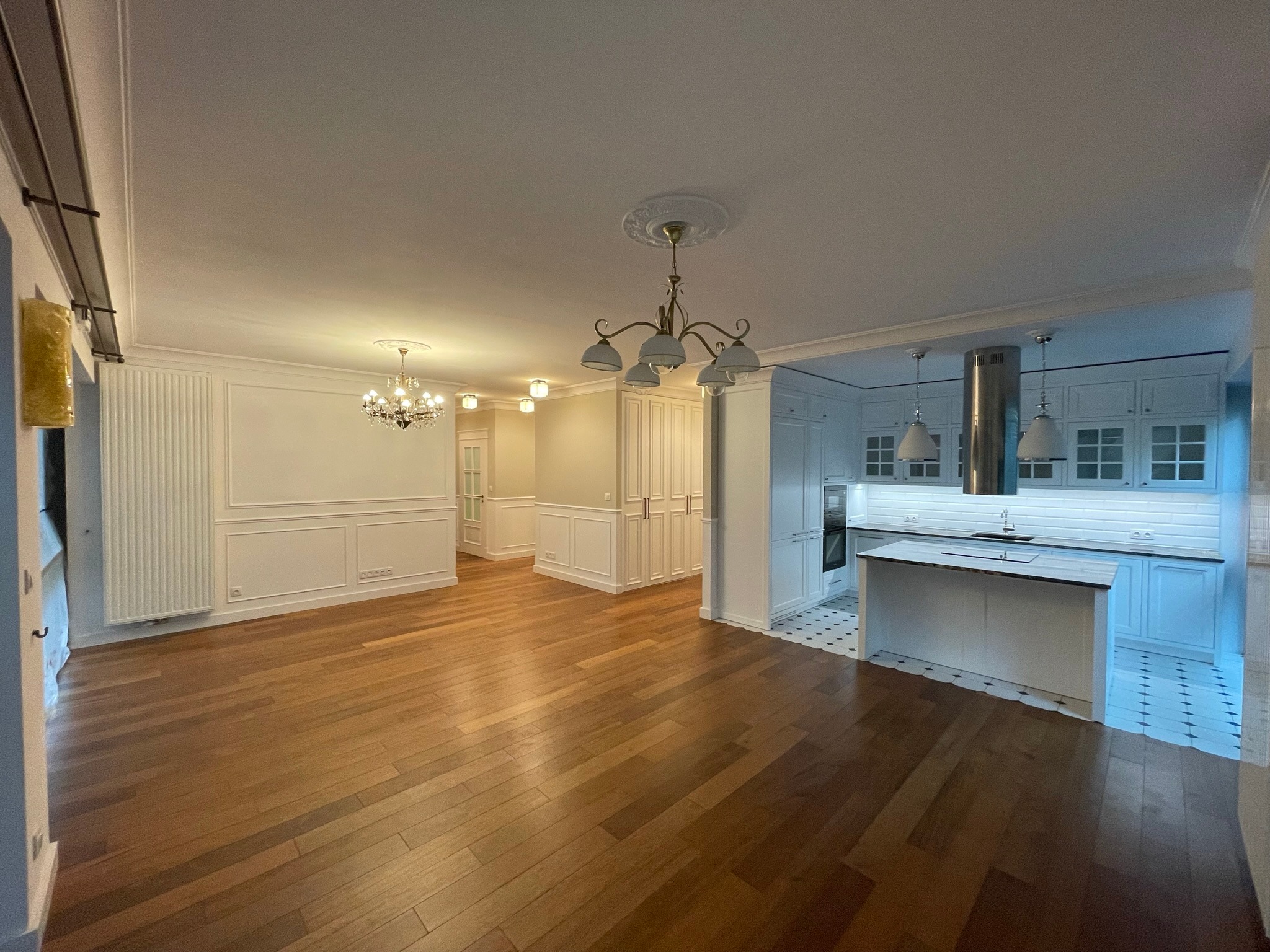 Przestronne wnętrze po remoncie: parkiet w jodełkę, białe ściany z dekoracyjnymi listwami, kuchnia z wyspą i białymi szafkami, żyrandole w klasycznym stylu.